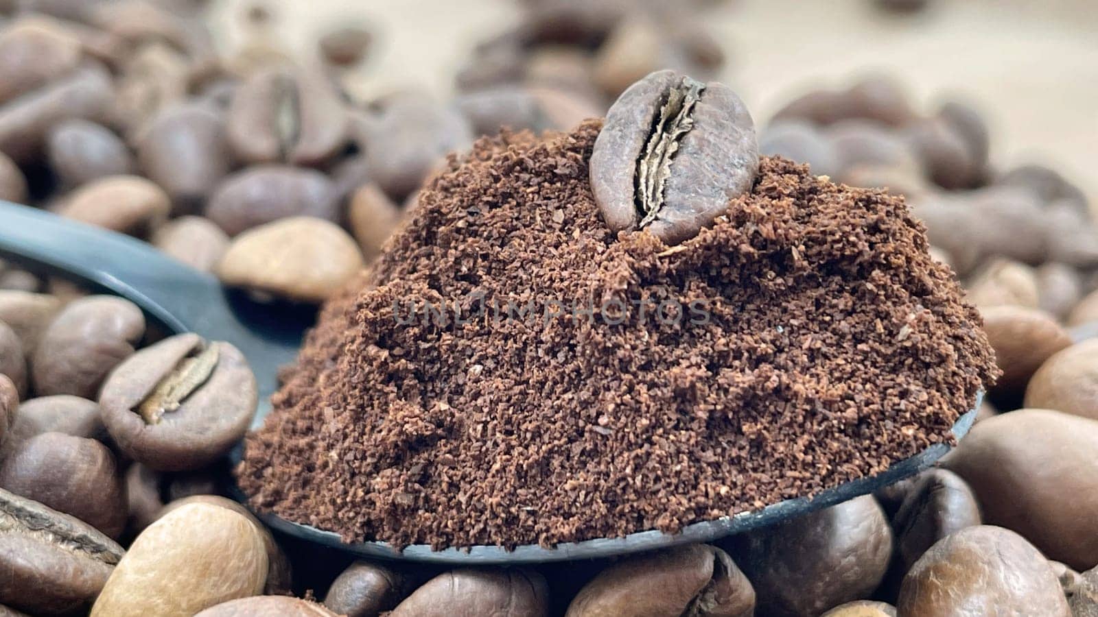 Against the background of roasted aromatic coffee beans lies a metal spoon filled with ground coffee. A drink made from roasted and ground beans from the coffee tree or coffee bush