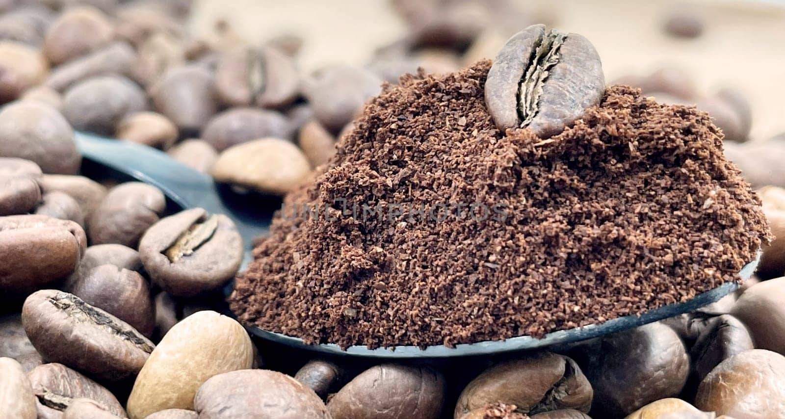 Against the background of roasted aromatic coffee beans lies a metal spoon filled with ground coffee. A drink made from roasted and ground beans from the coffee tree or coffee bush