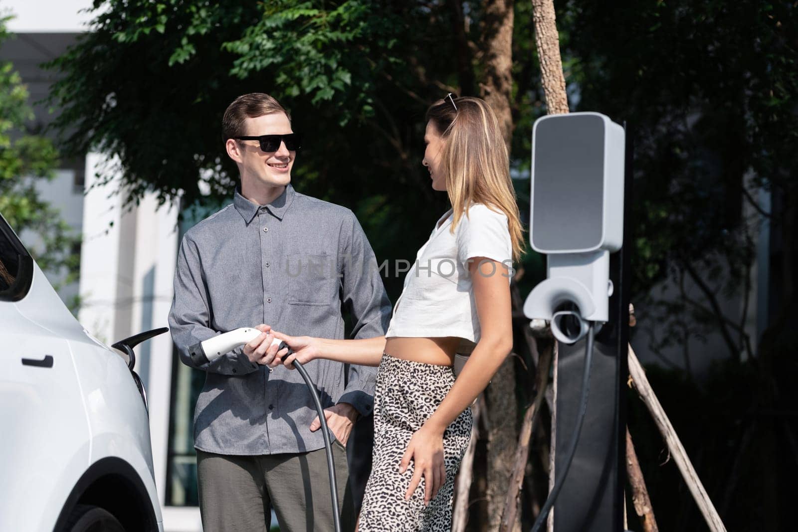 Young couple travel with EV electric car charging in green sustainable city outdoor garden in summer shows urban sustainability lifestyle by green clean rechargeable energy of electric vehicle innards