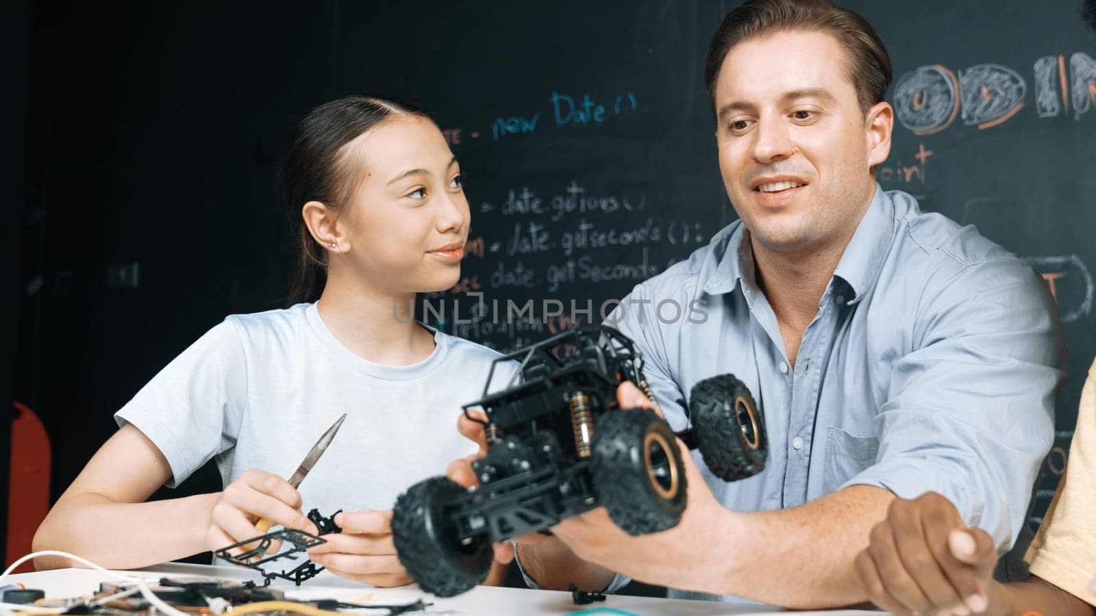 Smart teacher inspect car model construction to diverse student. Highschool children with mixed races listening mentor explain robotic model system at table with laptop and wires placed. Edification.