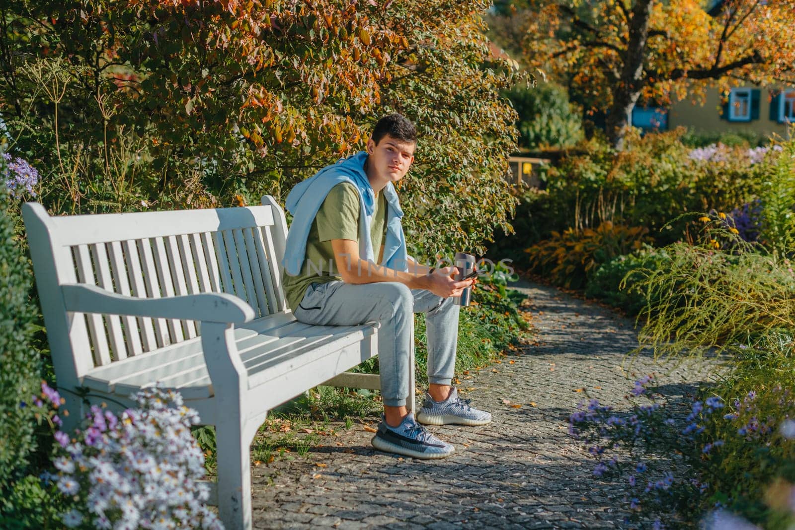 A Teenager Sits On A Bench In The Autumn Park Drinks Coffee From A Thermo Mug And Looks Into A Phone. Portrait Of Handsome Cheerful Guy Sitting On Bench Fresh Air Using Device Browsing Media Smm Drinking Latte Urban Outside Outdoor by Andrii_Ko