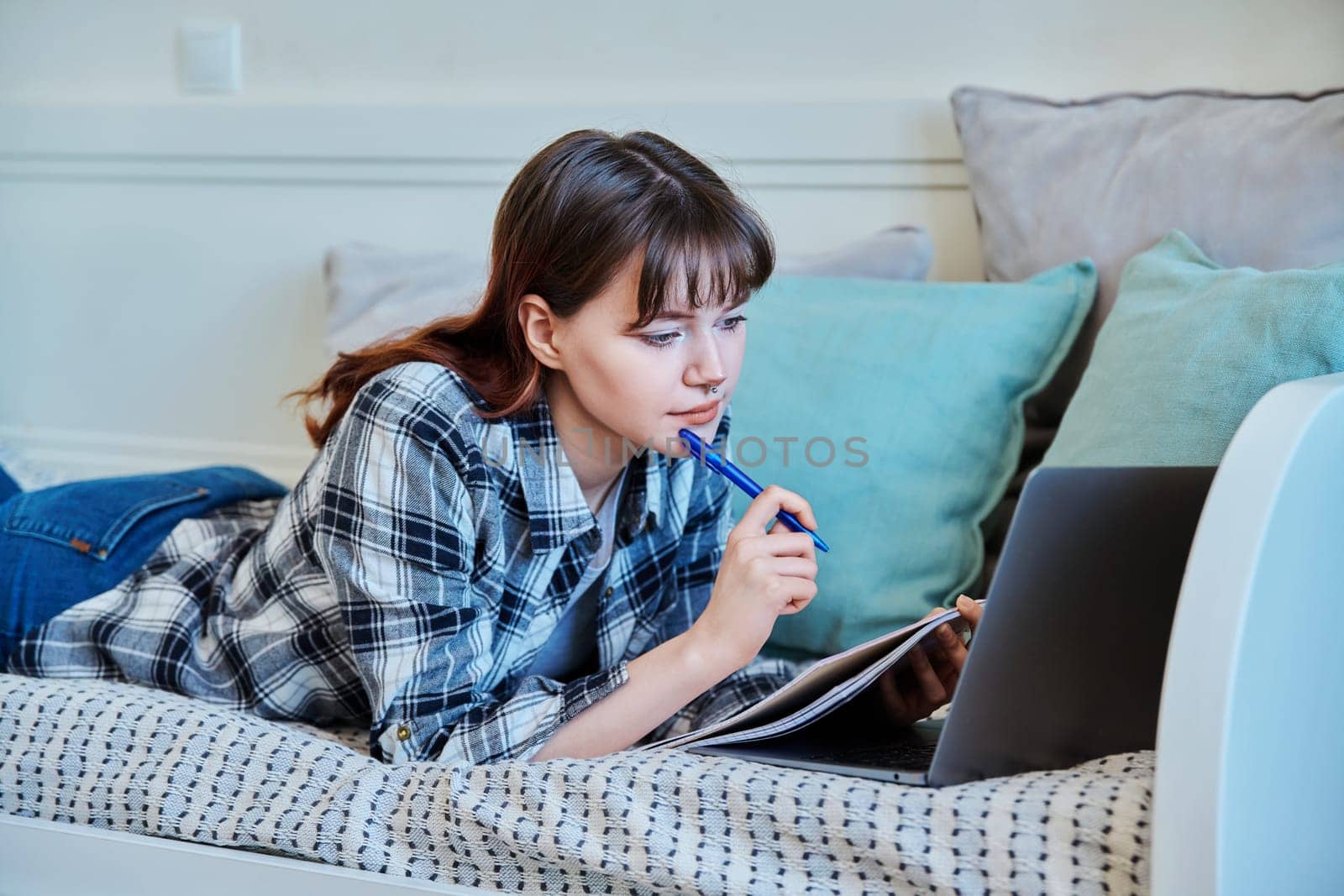 College student girl studying at home, lying on the couch with laptop, making notes in study book. Education, training, technology, youth concept