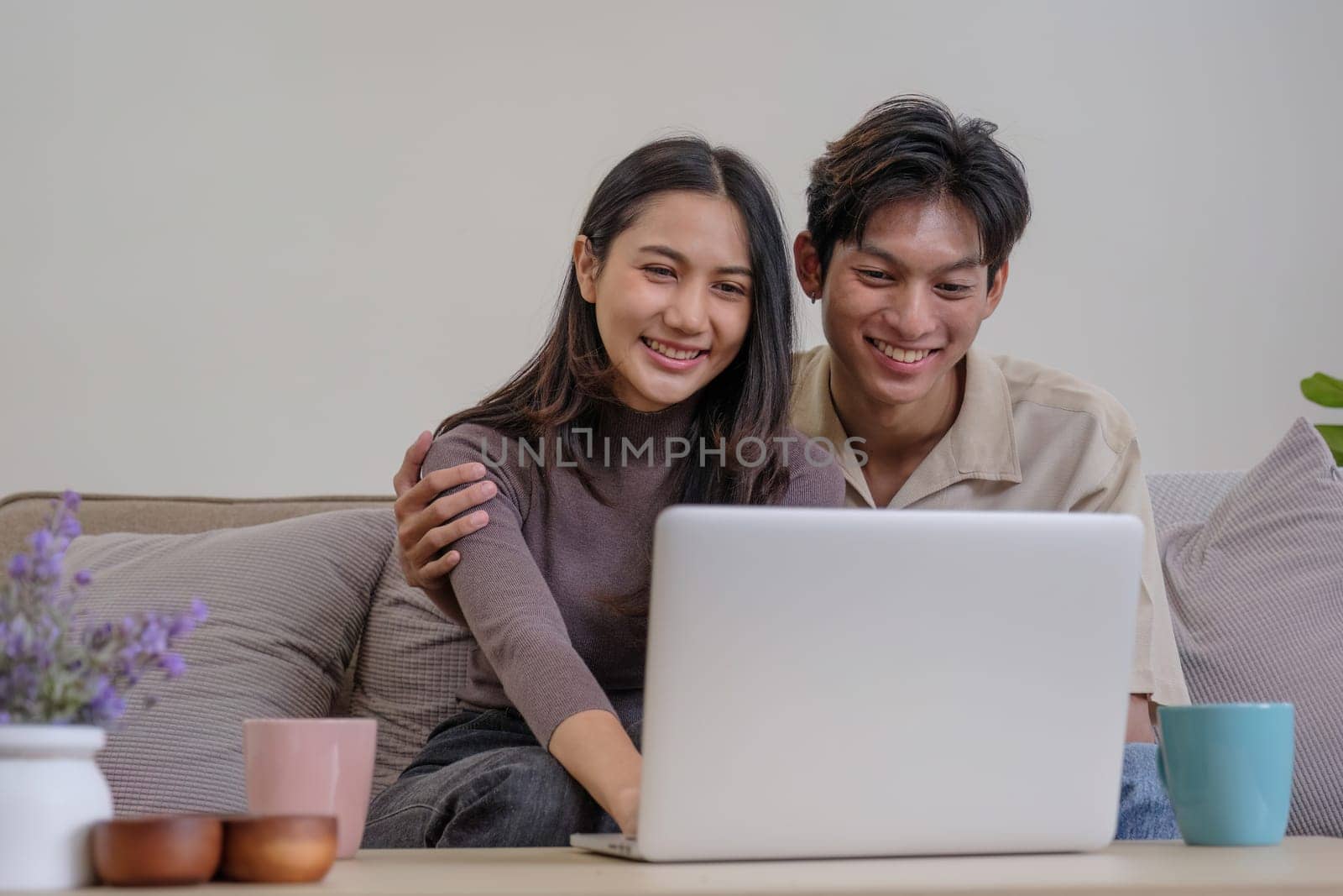 Asian couple watching movies on the internet using laptop at home Smiling Thai man and woman sitting on sofa, hugging each other, looking at computer screen. Surf the web together copy space by wichayada