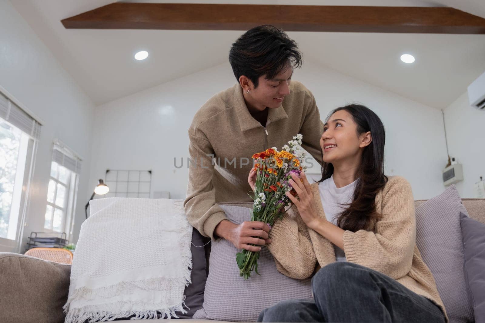 Romantic young asian couple embracing with holding flowers and smiling in living room at home. fall in love. Valentine concept.