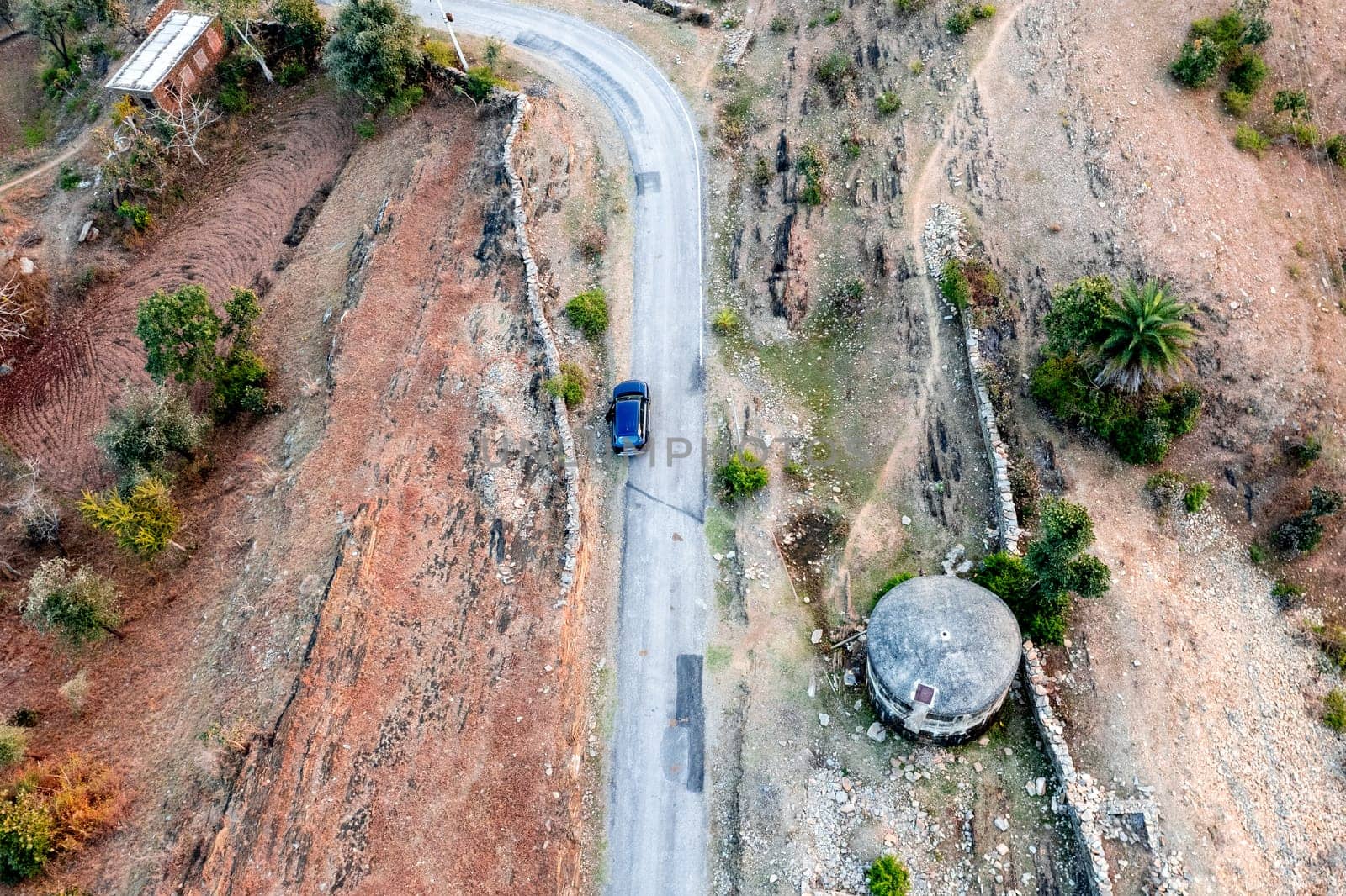 Aerial drone shot showing blue car moving on narrow country rural road with barren land on the side with green trees showing village town roads by Shalinimathur