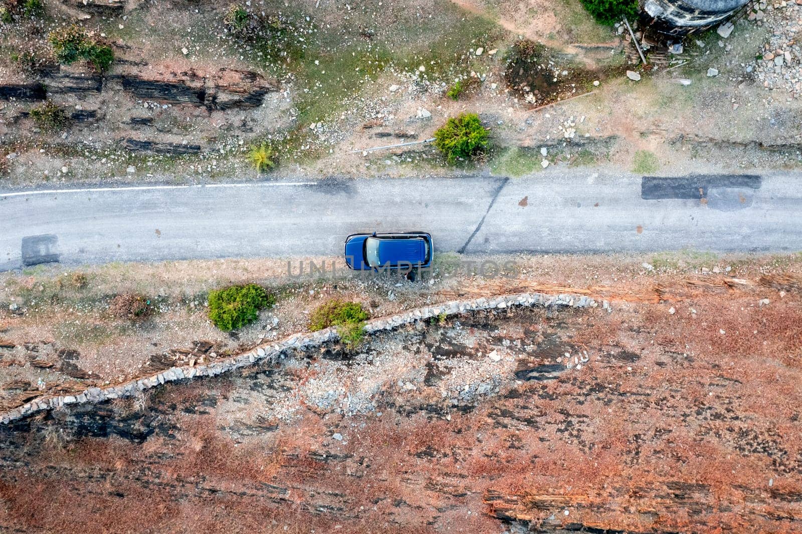 Aerial drone shot showing blue car moving on narrow country rural road with barren land on the side with green trees showing village town roads Udaipur India