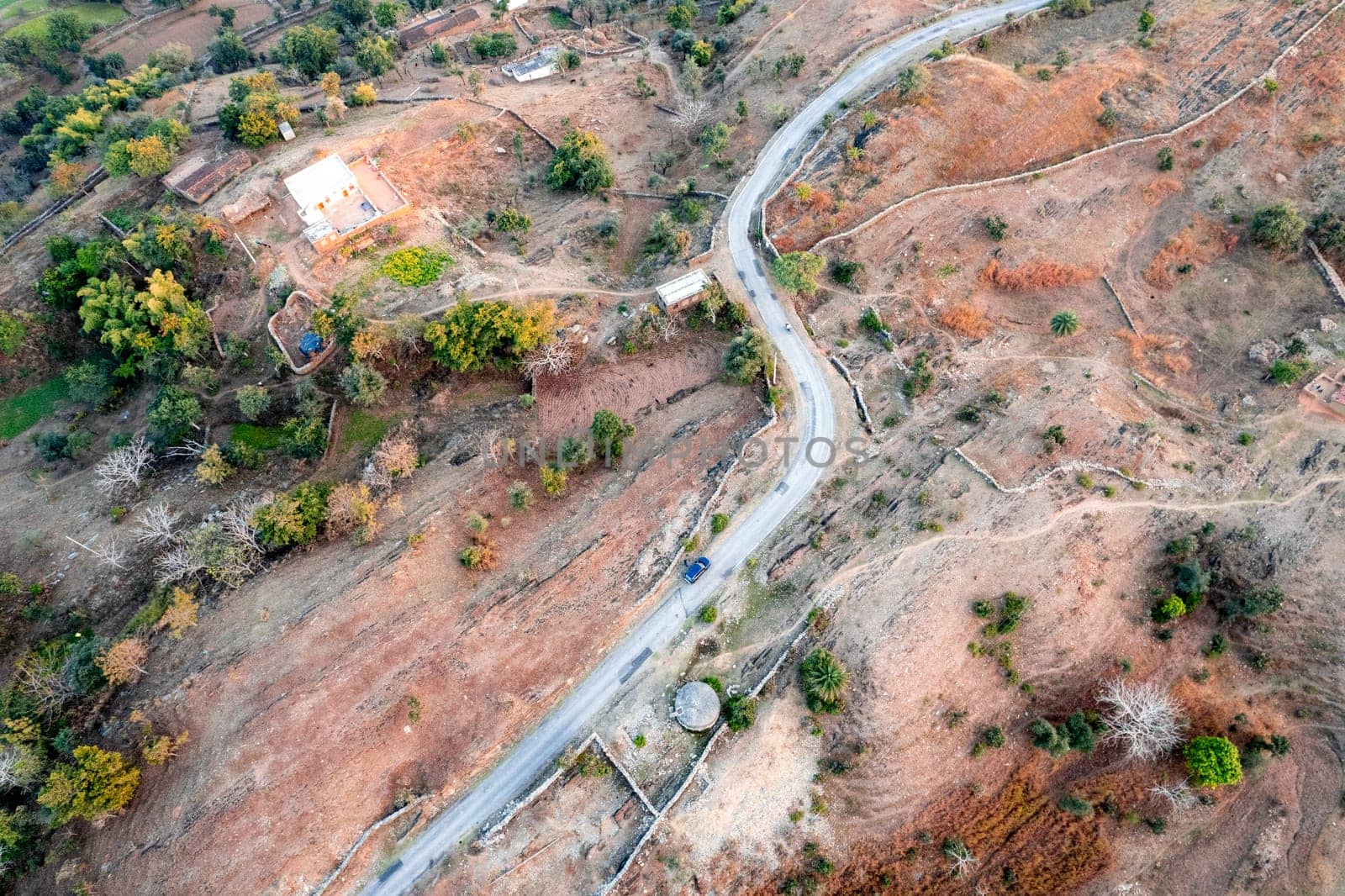 Aerial drone shot showing blue car moving on narrow country rural road with barren land on the side with green trees showing village town roads by Shalinimathur