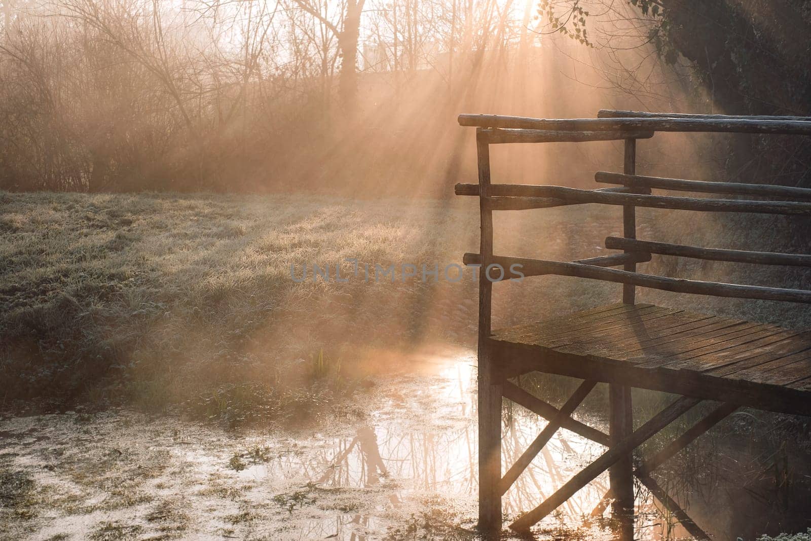 Wooden bridge over lake in early misty morning, fog over the water on sun rays.