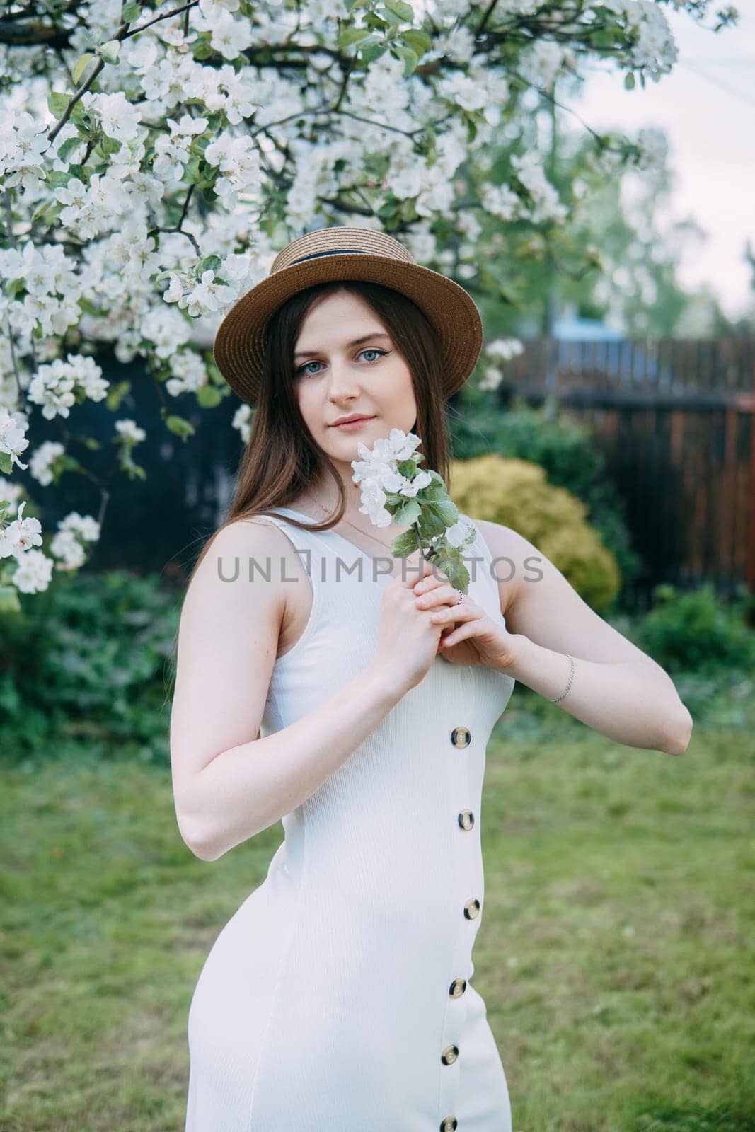 Beautiful young girl in white dress and hat in blooming Apple orchard. Blooming Apple trees with white flowers