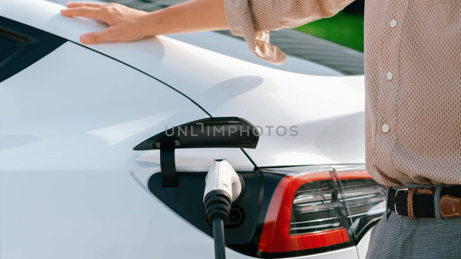 Young man put EV charger to recharge electric car's battery from charging station in city commercial parking lot. Rechargeable EV car for sustainable environmental friendly urban travel. Expedient