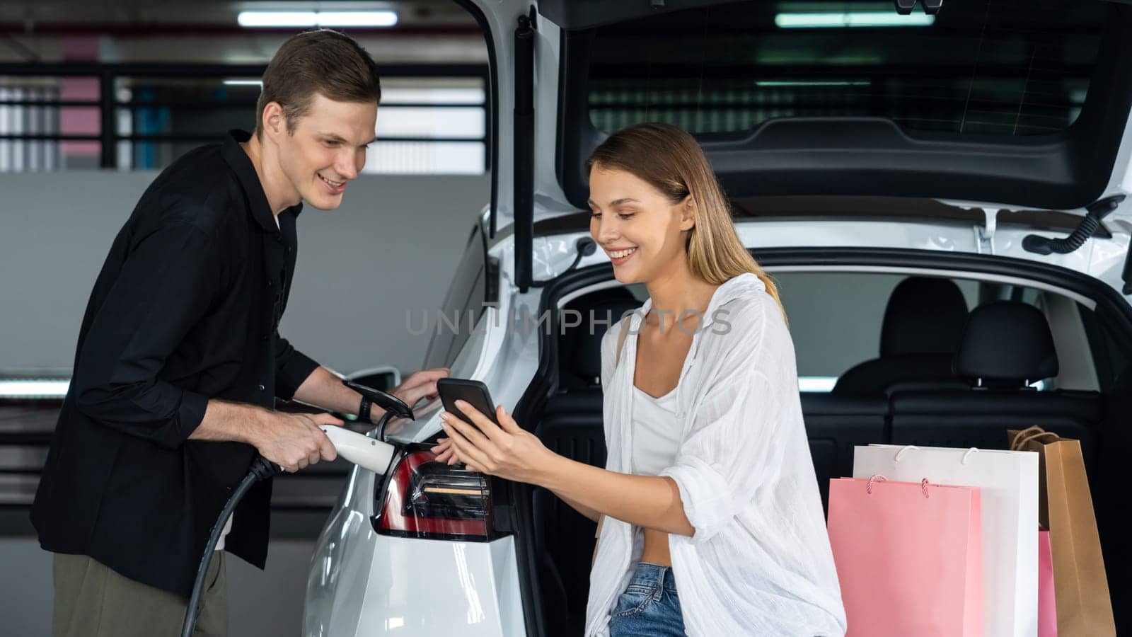 Young couple travel with EV electric car to shopping center parking lot charging in downtown city showing urban sustainability lifestyle by green clean rechargeable energy of electric vehicle innards