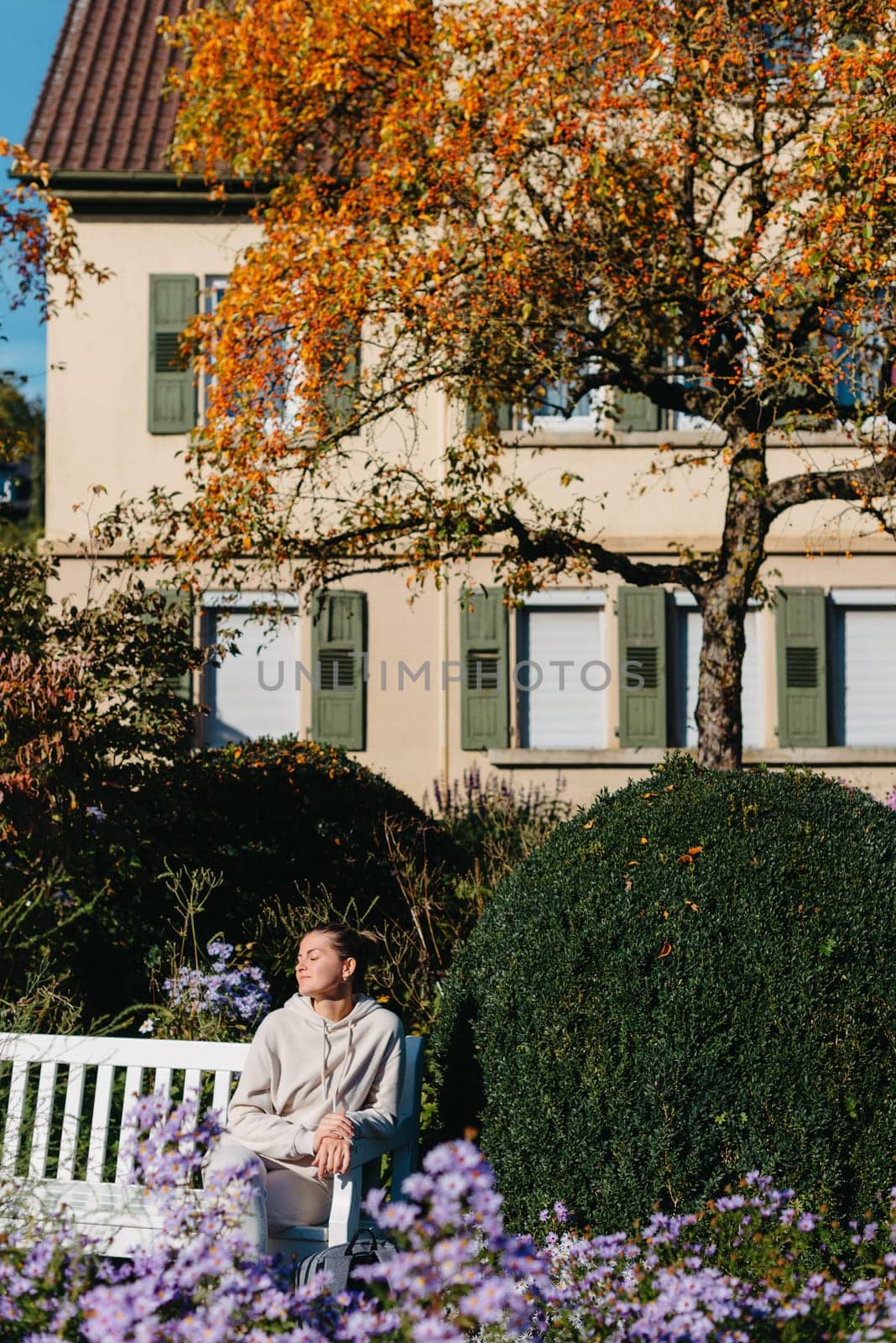 a girl sits on a bench in the park and enjoys the sun. Portrait young adult attractive woman enjoy sitting on bench and relaxing calm carefree rest in city park against green grass and trees on sunny day. Single female person relaxing chilling outdoors