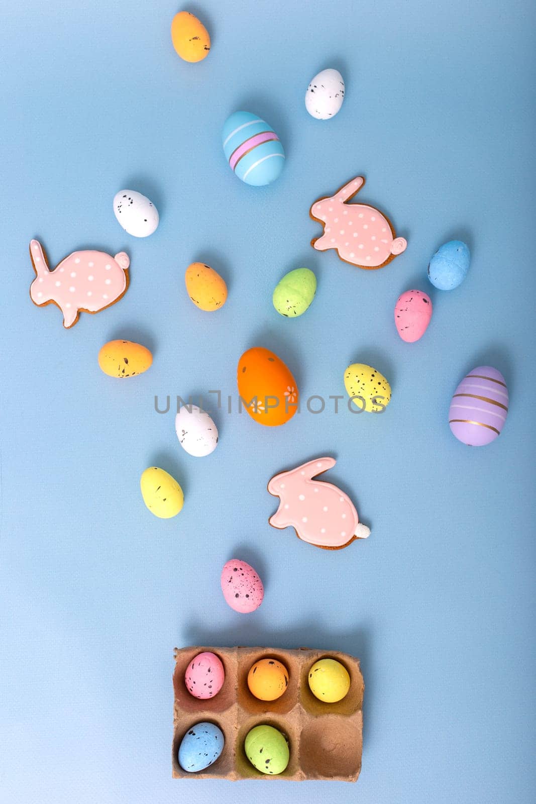 Beautiful decorative multicolored eggs and pink gingerbread in the form of rabbit, scattered on a light blue background, top view. Vertical
