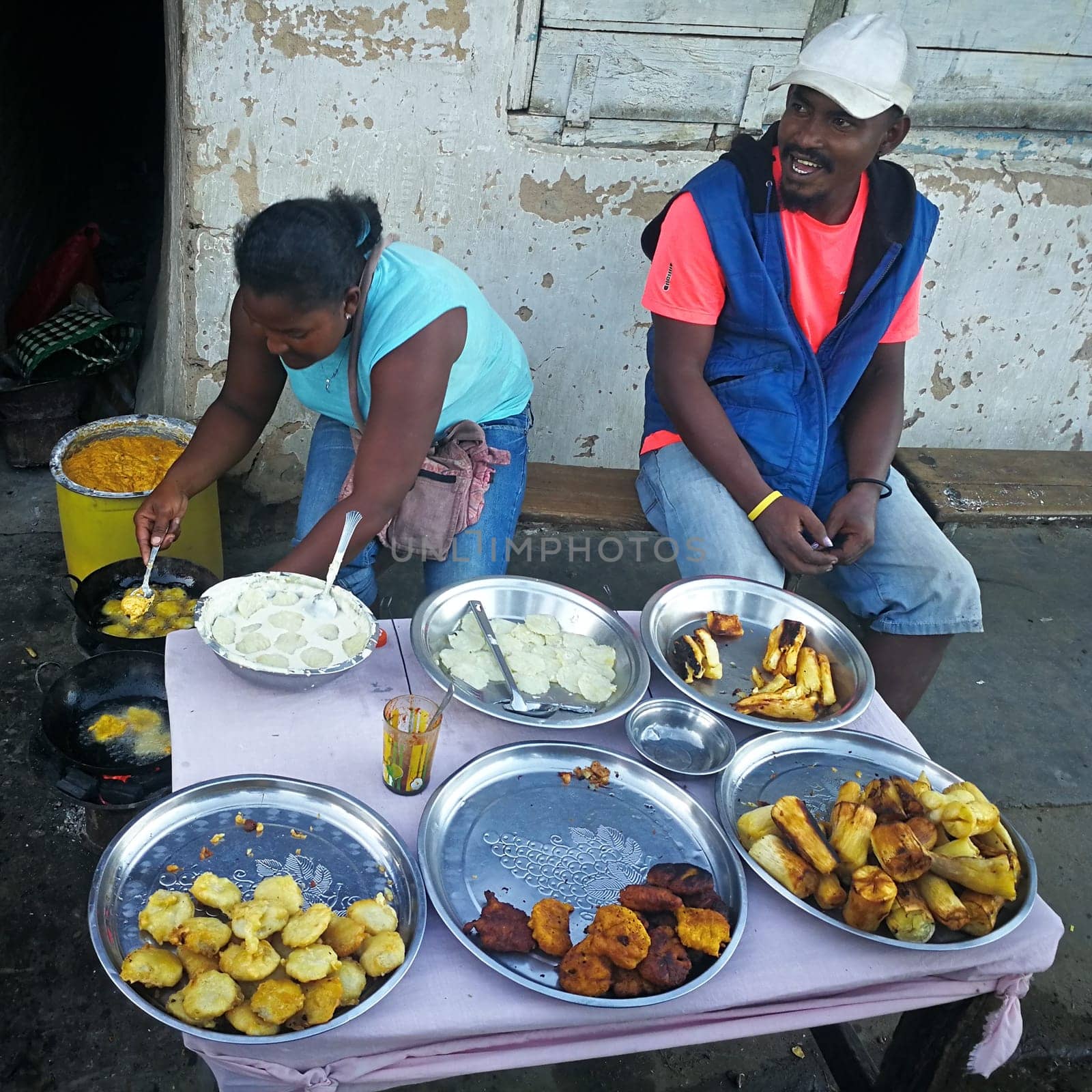 Ranohira, Madagascar - April 29, 2019: Unknown Malagasy woman cooks on the street, deep frying various coated vegetables, another man sits next to her on wooden bench. Food is often cooked at streets by Ivanko