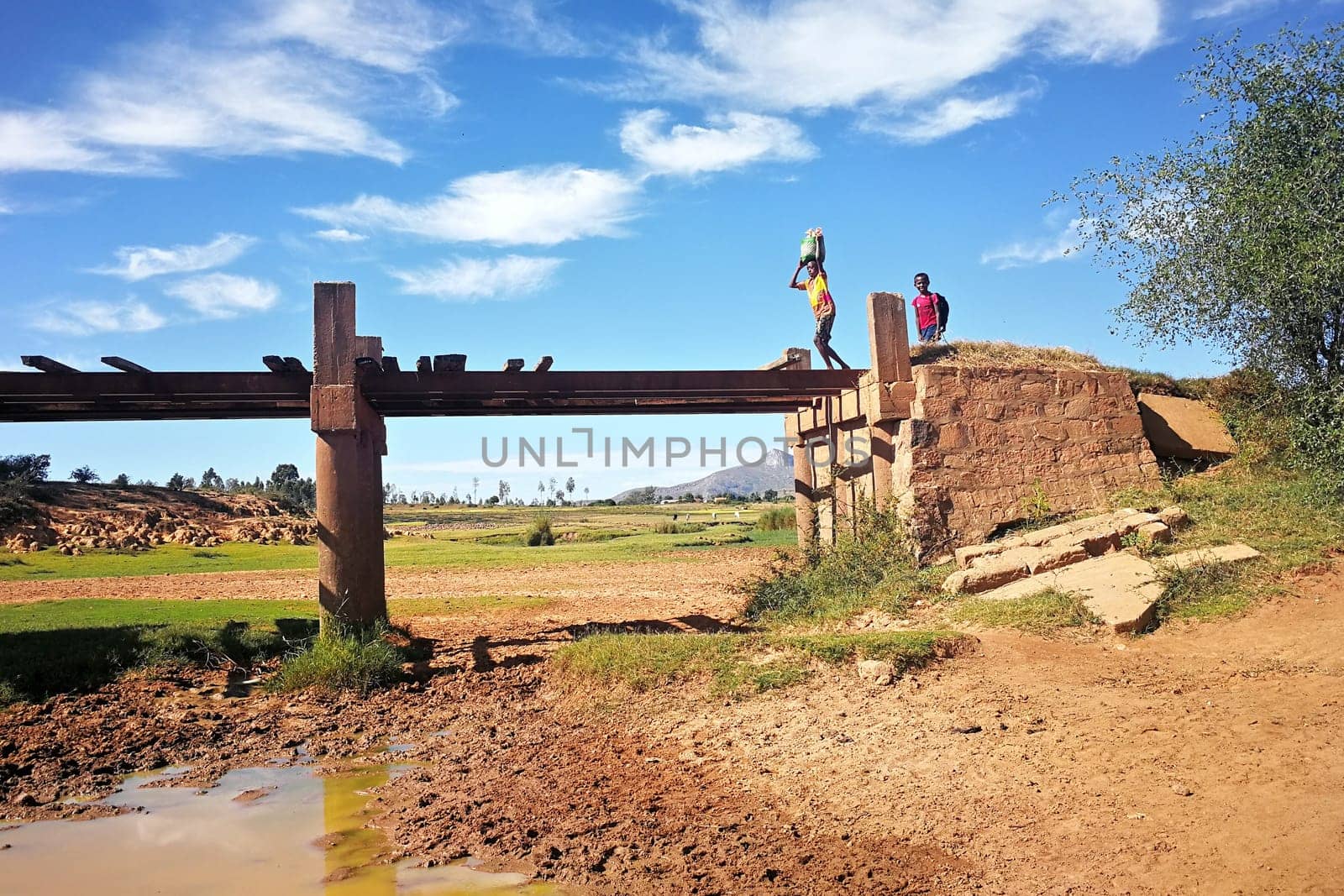 Ambalavao, Madagascar - April 27, 2019: Unknown Malagasy boy carrying bag on his head over simple bridge, another one smiling behind him, typical Madagascar landscape on sunny day in background by Ivanko