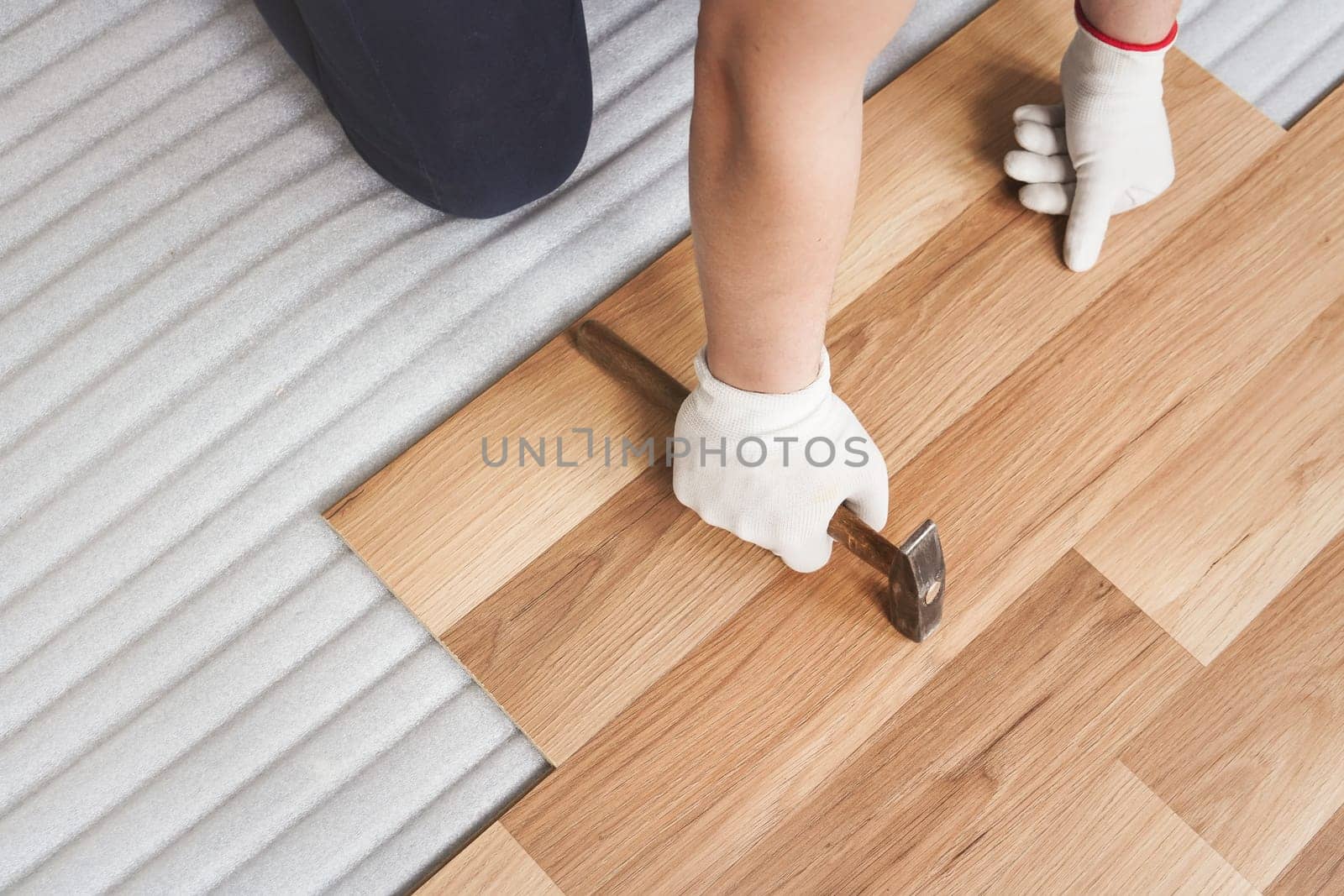 Installing laminated floor, detail on man hands holding in white gloves, hammer in one hand, leaning over wooden tiles, white foam base layer under by Ivanko