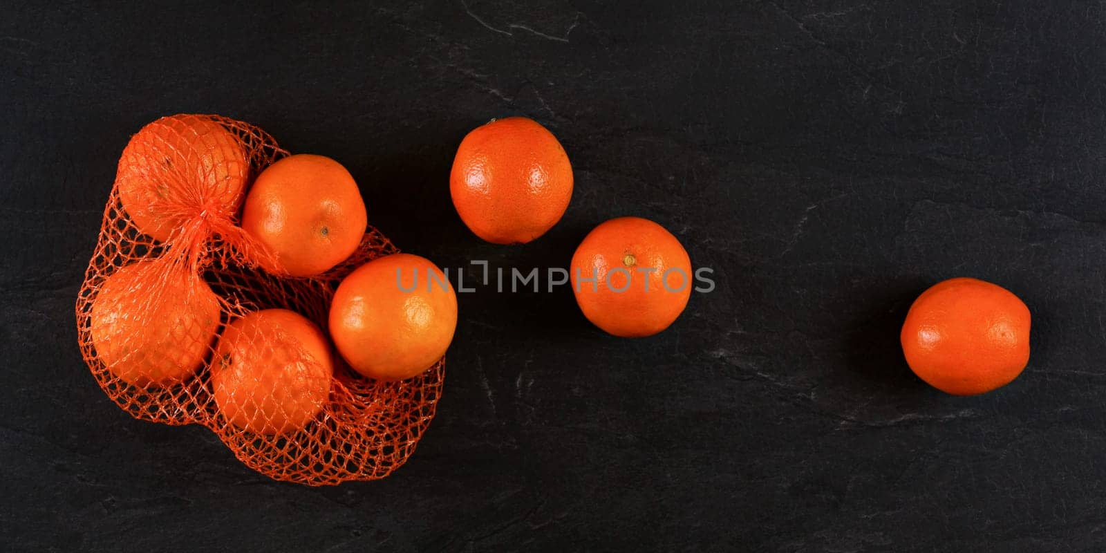 Oranges in plastic red net from supermarket, some scattered on black stone like board, photo from above by Ivanko