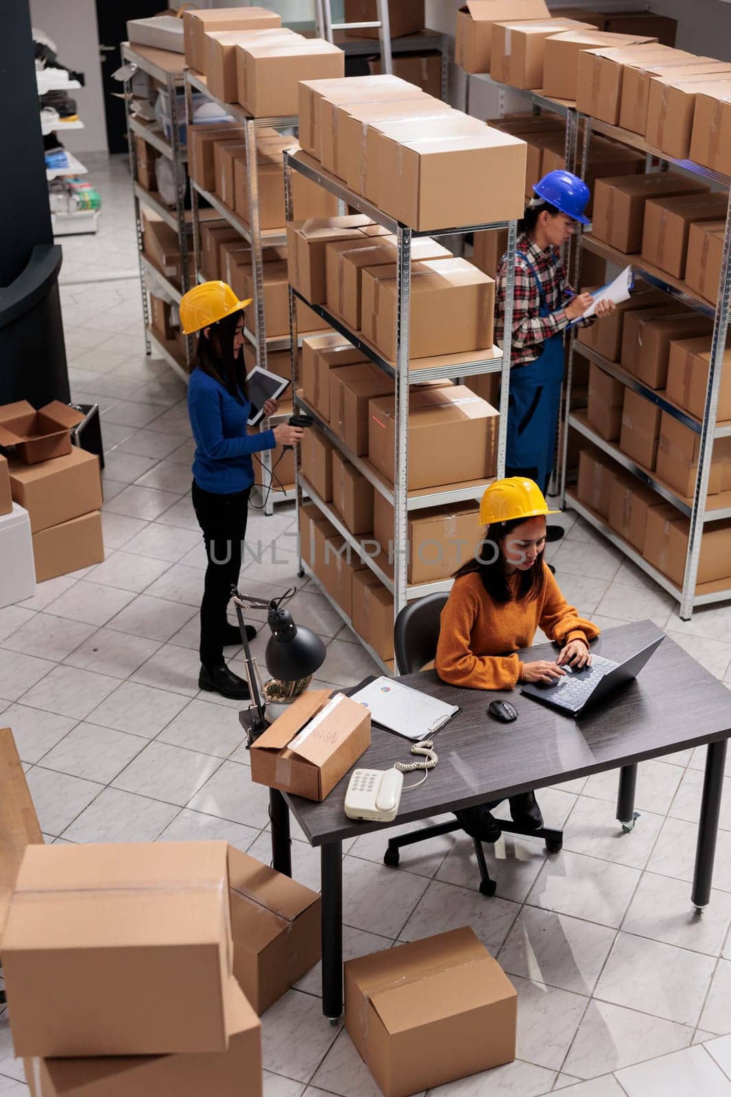 Industrial storehouse asian coworkers doing inventory in storage room full of cardboard boxes on shelves. Postal service warehouse young colleagues team in protective helmets working together top view