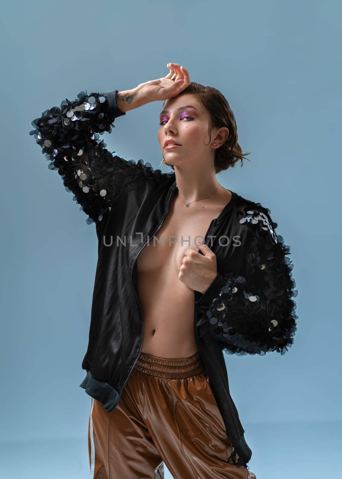 Portrait of sensual young female model with short hair in black fashionable jacket. Sexy woman with makeup in trendy outfit on naked body against blue background.