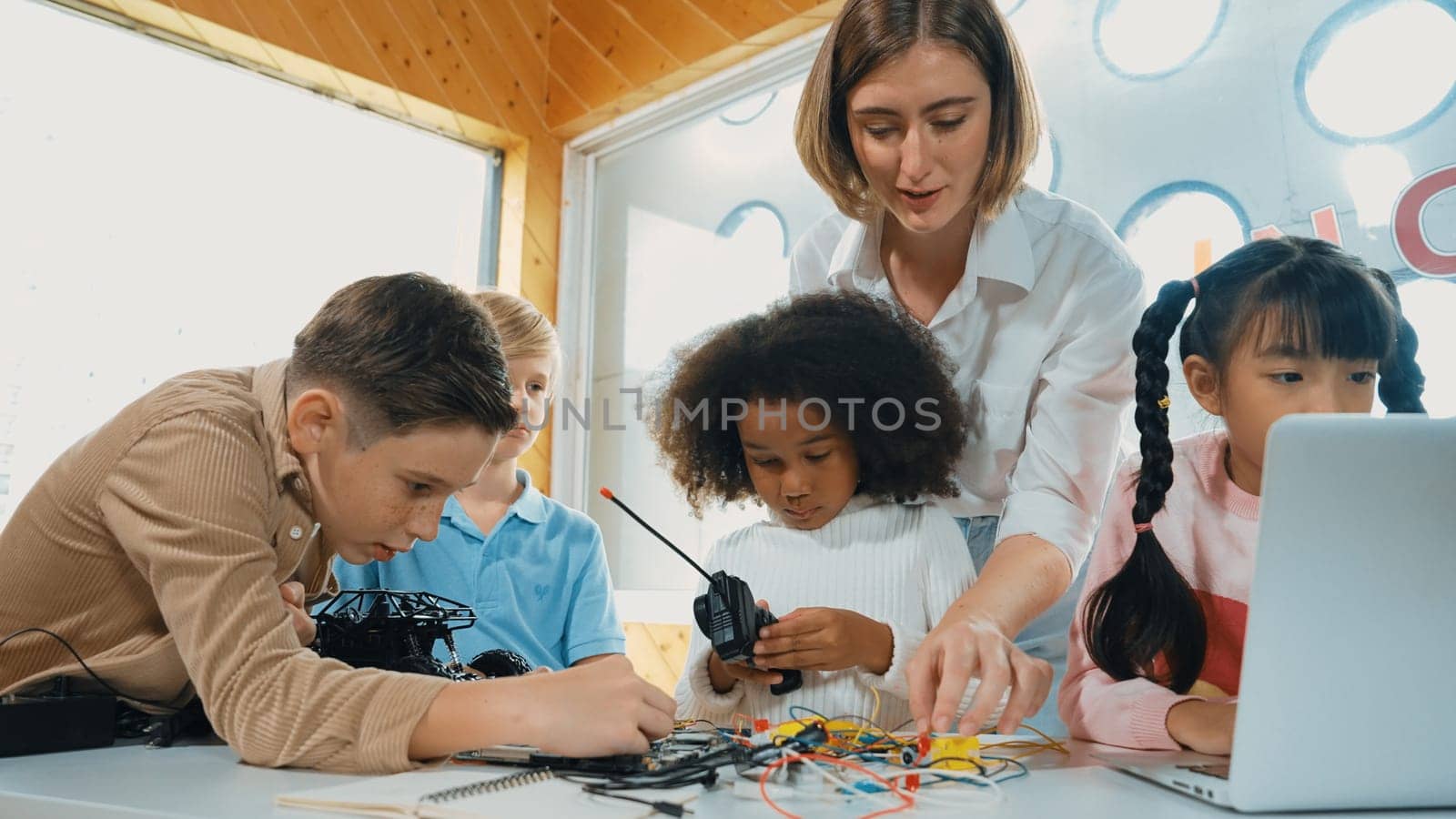 Teacher teach and explain students about digital electrical tool or car model. Diverse kid learn electronic equipment and use to fix motherboard or robotic model at table with wires placed. Erudition.