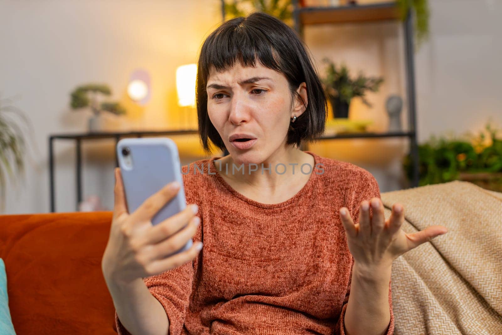 Upset displeased woman with short hair using smartphone sitting on sofa in living room at home. Unhappy Hispanic girl in casual clothes loses becoming surprised by sudden lottery results or bad news.
