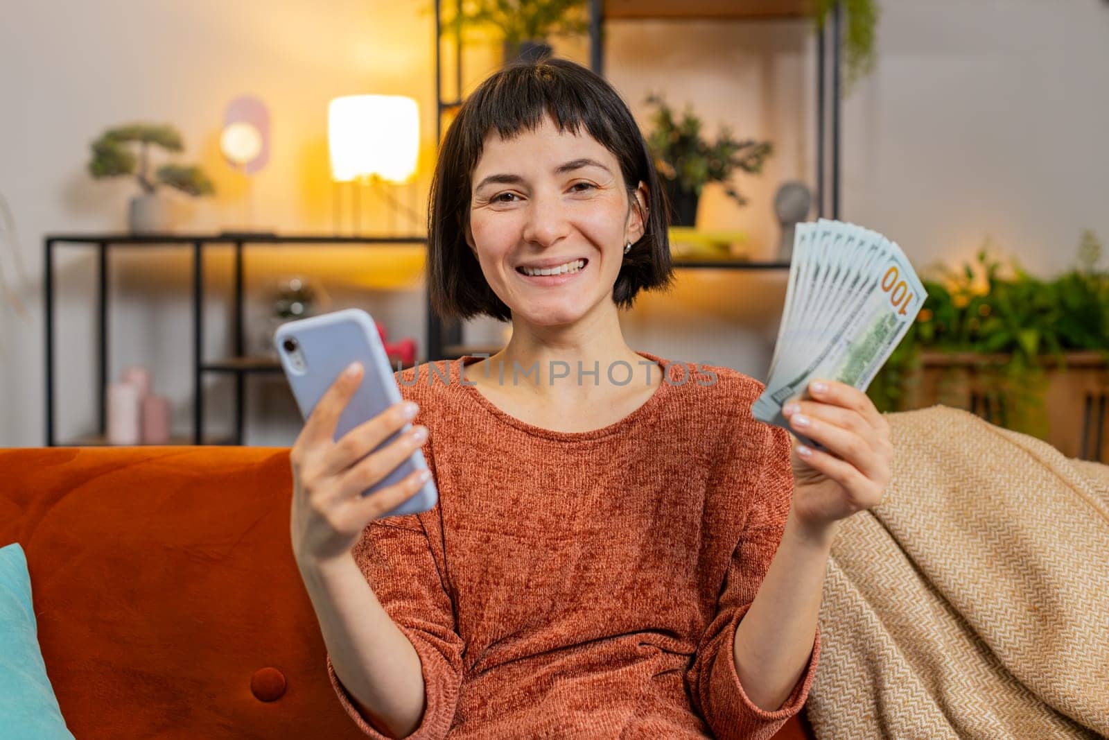 Smiling happy woman counting money dollar banknotes using smartphone calculator on sofa in room at home. Hispanic lovely girl calculating budget, savings, earnings and planning vacation in apartment