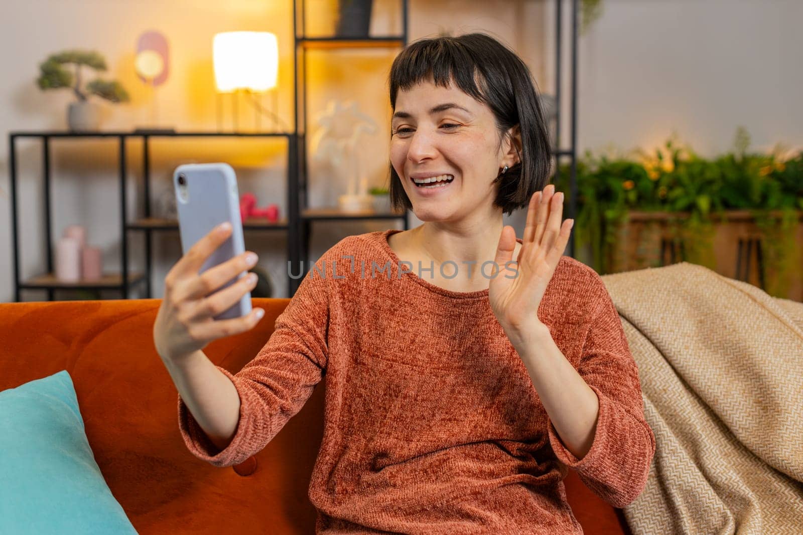 Smiling young woman in casual clothes waving on video call via smartphone in apartment. Happy Caucasian girl having conversation with friend on mobile phone sitting on sofa in living room at home.