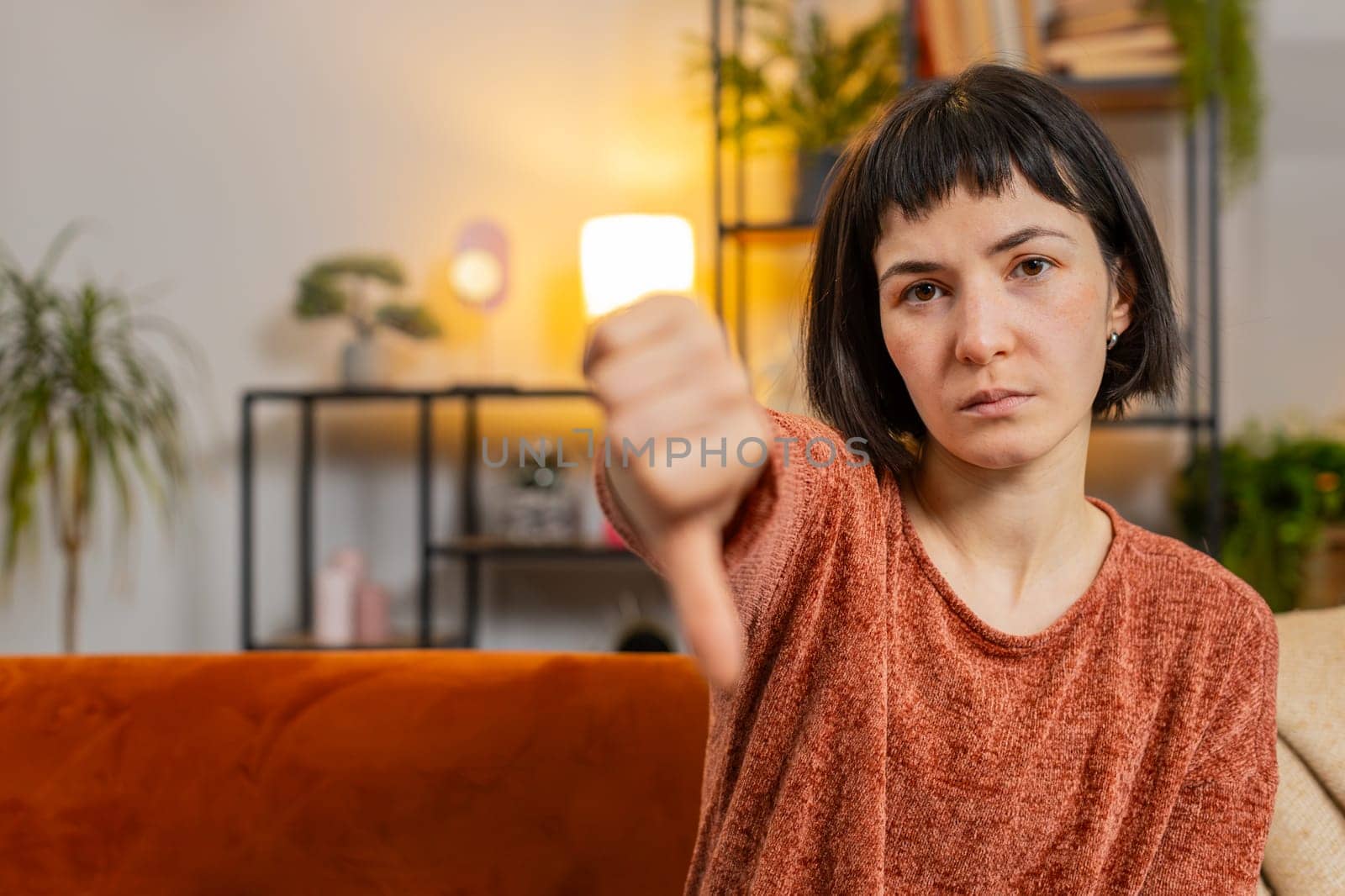 Portrait of upset Caucasian woman showing double thumbs down sign gesture, expressing discontent disapproval dissatisfied bad work dislike sitting in living room at home. Girl sits on sofa. Copy-space