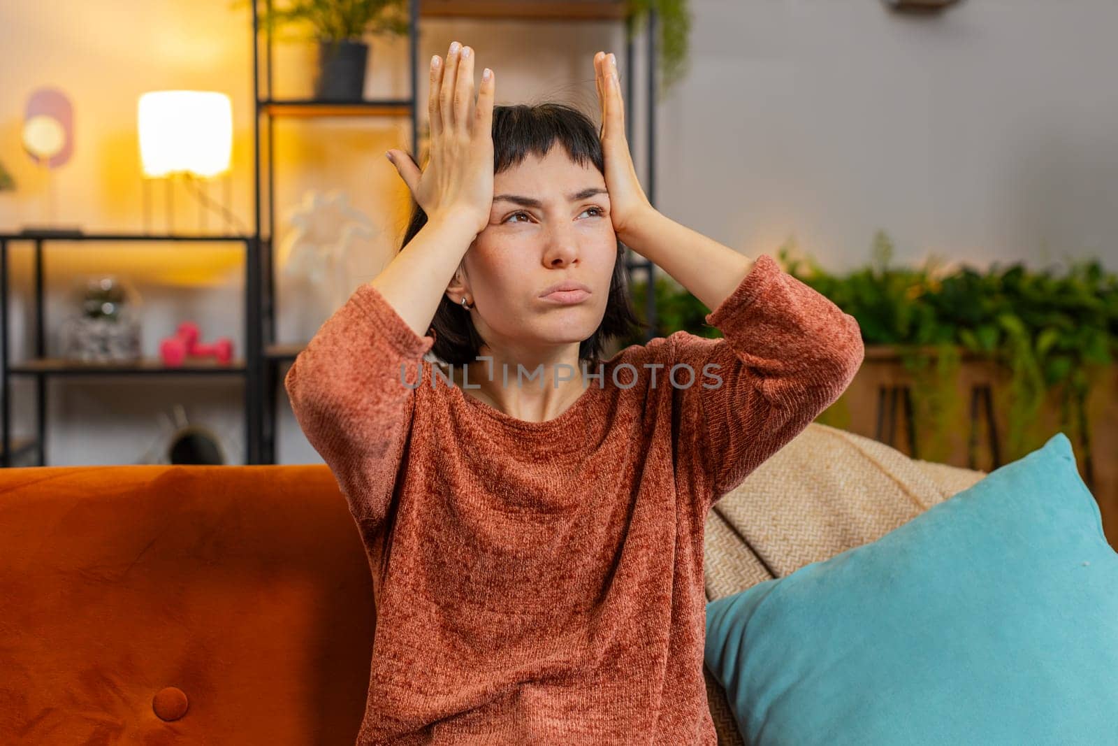 Displeased Caucasian young woman rubbing temples to cure headache problem, suffering from tension and migraine, stress grimacing in pain. Upset girl sitting on couch in living room at home apartment