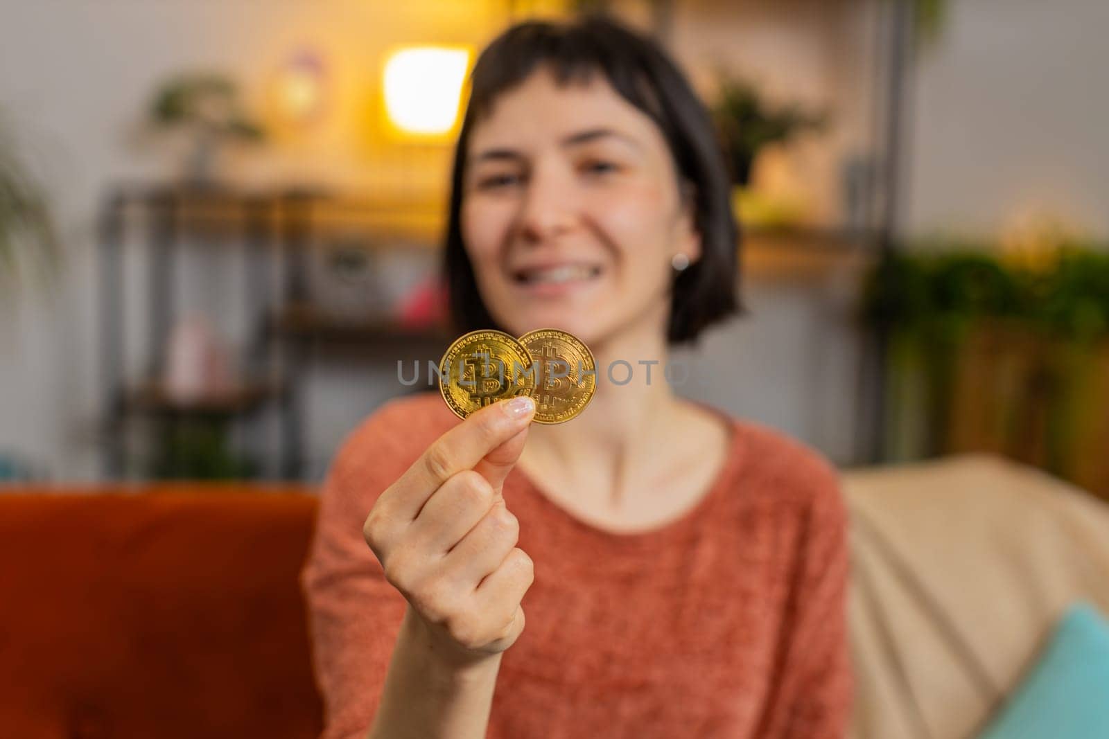 Portrait of smiling Caucasian woman holding two golden bitcoins BTC, looking at camera. Happy surprised girl stock trader showing thumbs up sitting on sofa in living room at home. Increasing wealth.