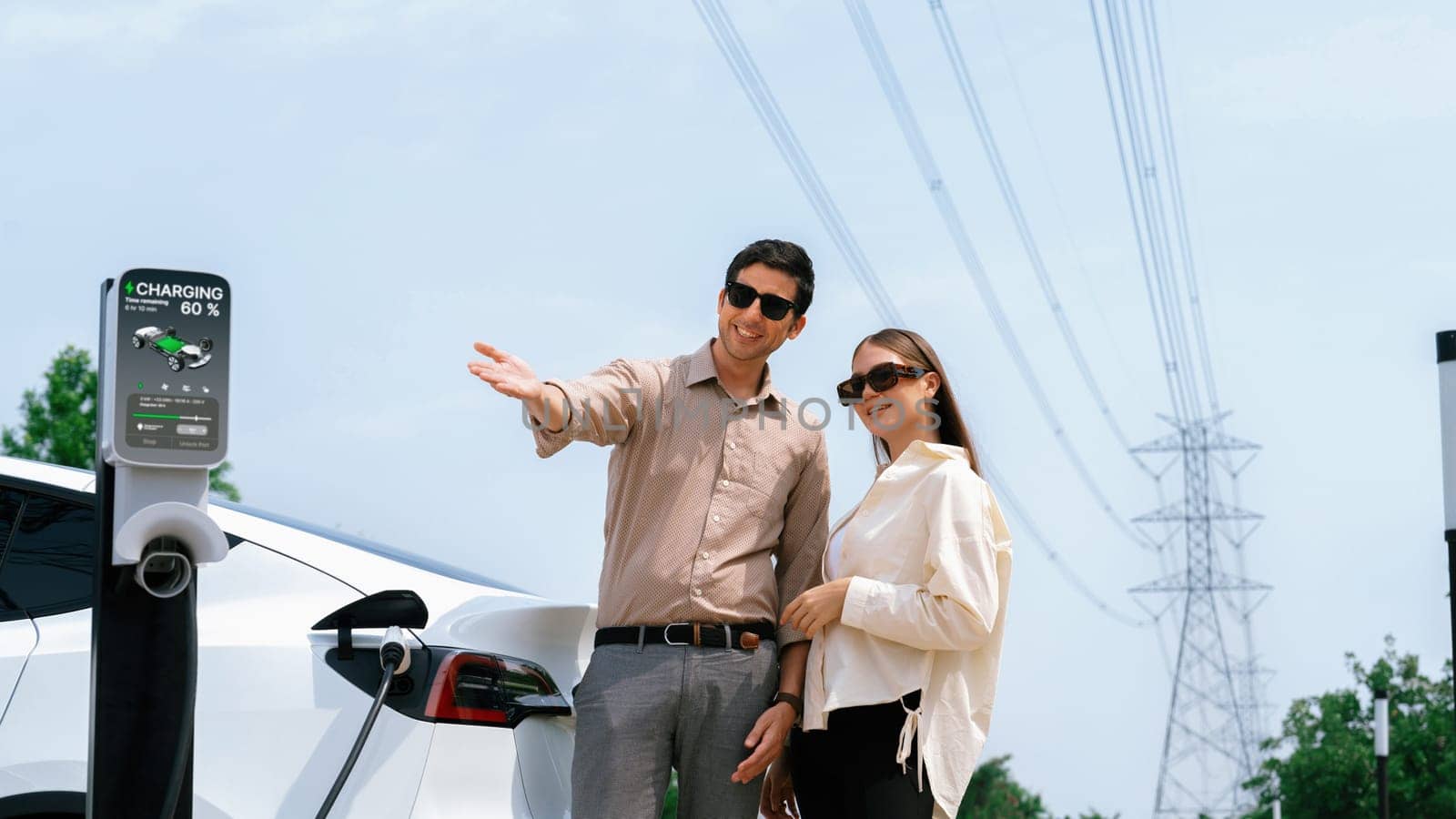 Young couple recharge EV car battery at charging station connected to power grid tower electrical industrial facility as electrical industry for eco friendly vehicle utilization. Expedient