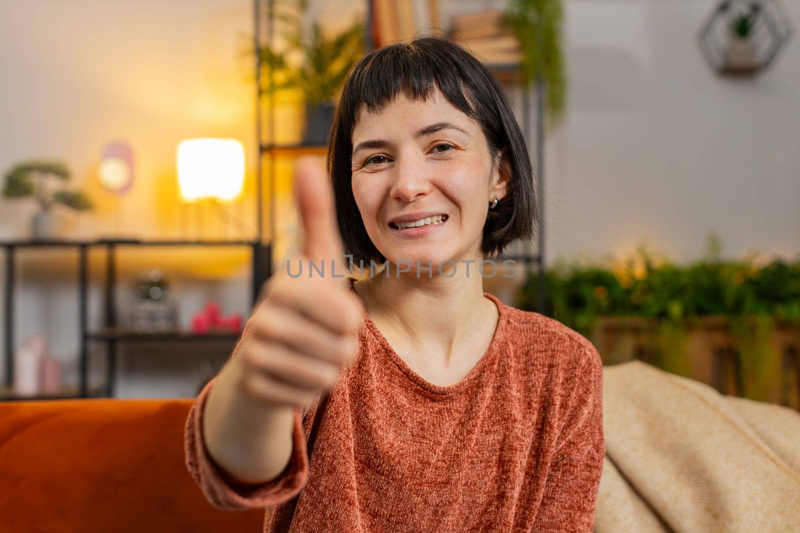 Portrait of excited Caucasian woman looking approvingly at camera showing double thumbs up, like sign positive, good news, positive feedback. Smiling girl sitting on couch in living room at home.