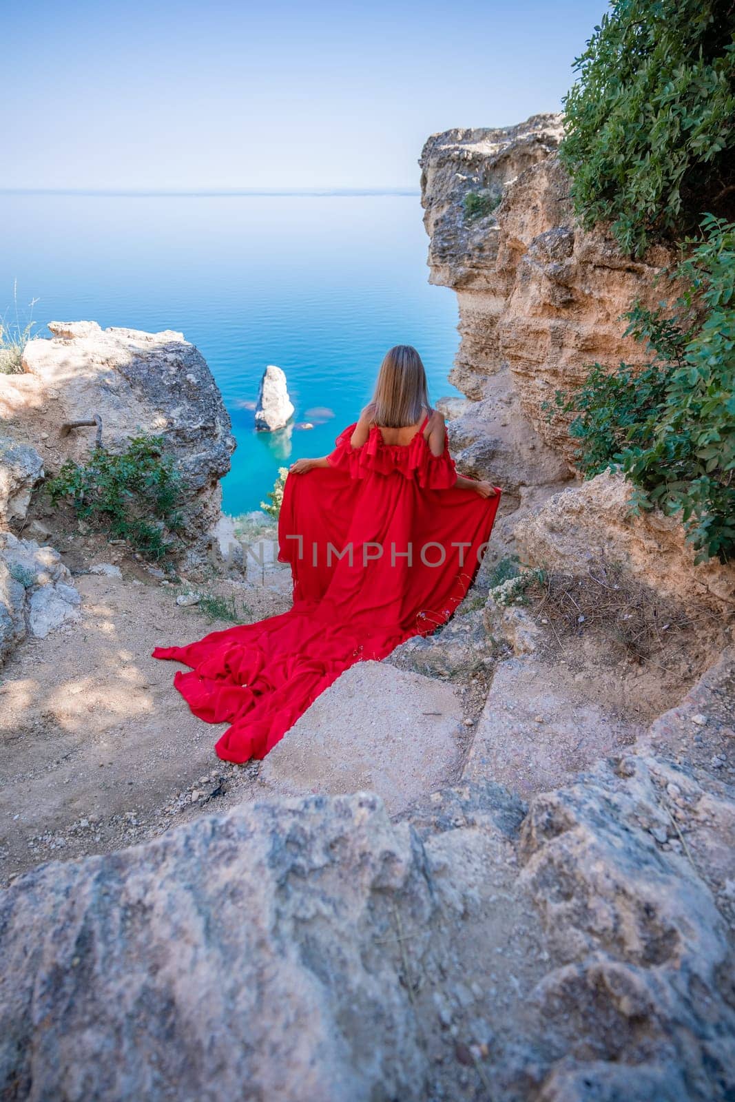 Red dress sea woman. Happy woman with flowing hair in a long flowing red dress stands on a rock near the sea. Travel concept, photo session at sea by Matiunina