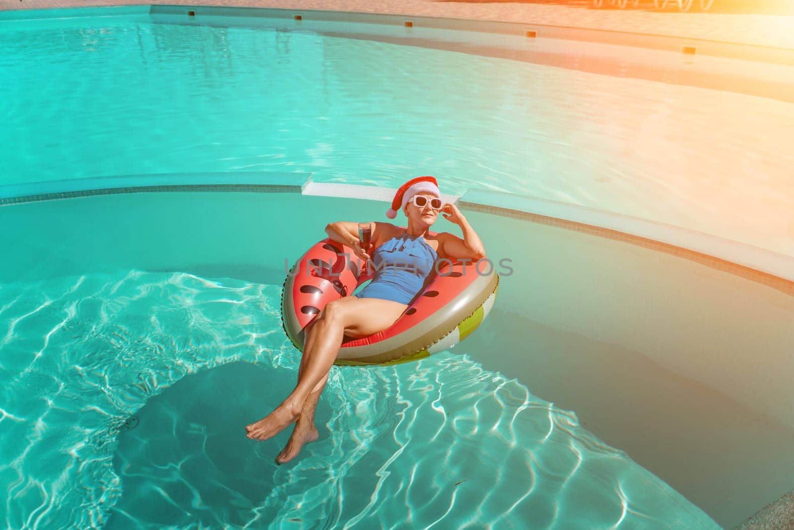 A happy woman in a blue bikini, a red and white Santa hat and sunglasses poses in the pool in an inflatable circle with a watermelon pattern, holding a glass of wine in her hands. Christmas holidays concept