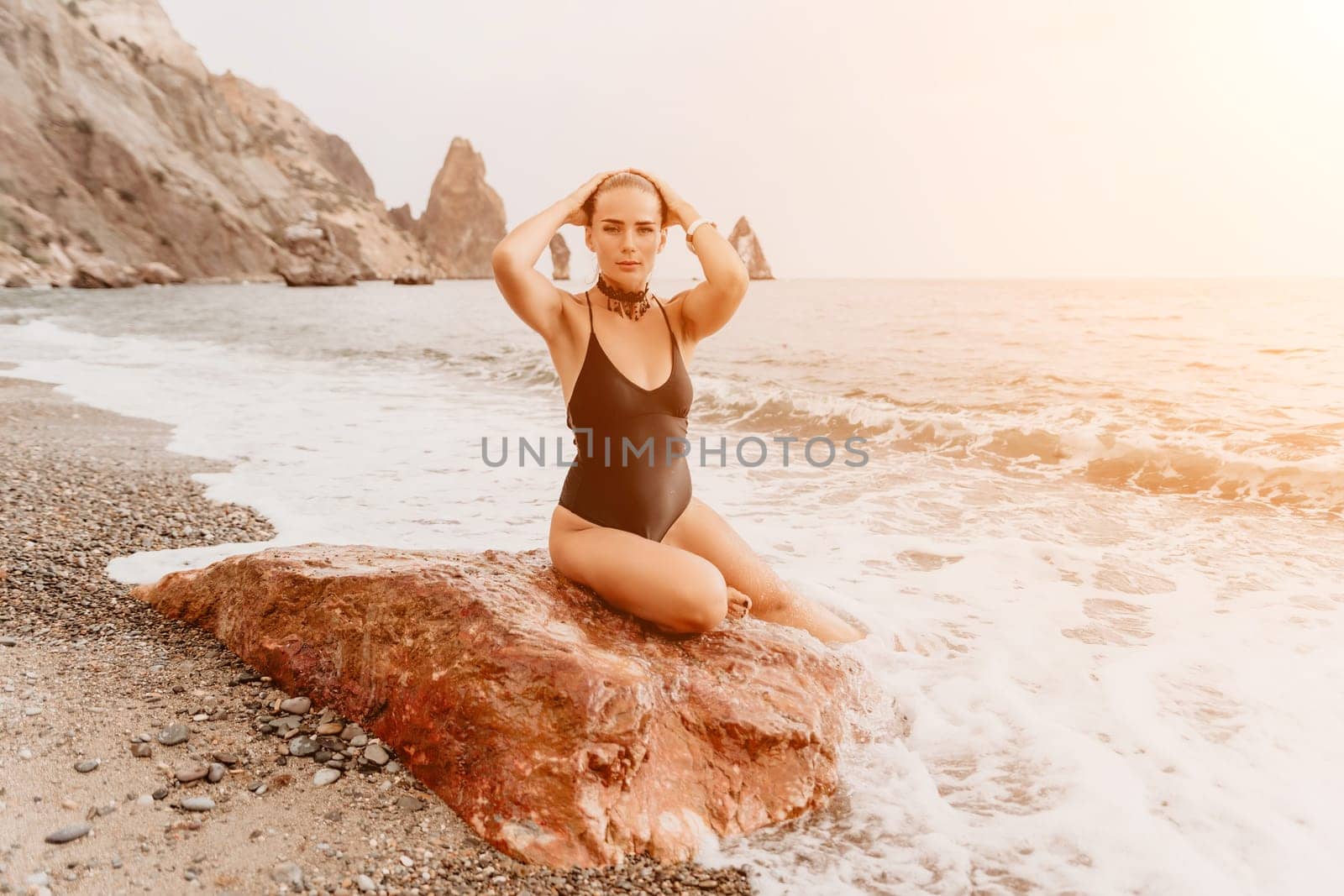 Woman travel portrait. Happy woman with long hair looking at camera and smiling. Close up portrait cute woman in black bikini posing on a red volcanic rock on the sea beach by panophotograph