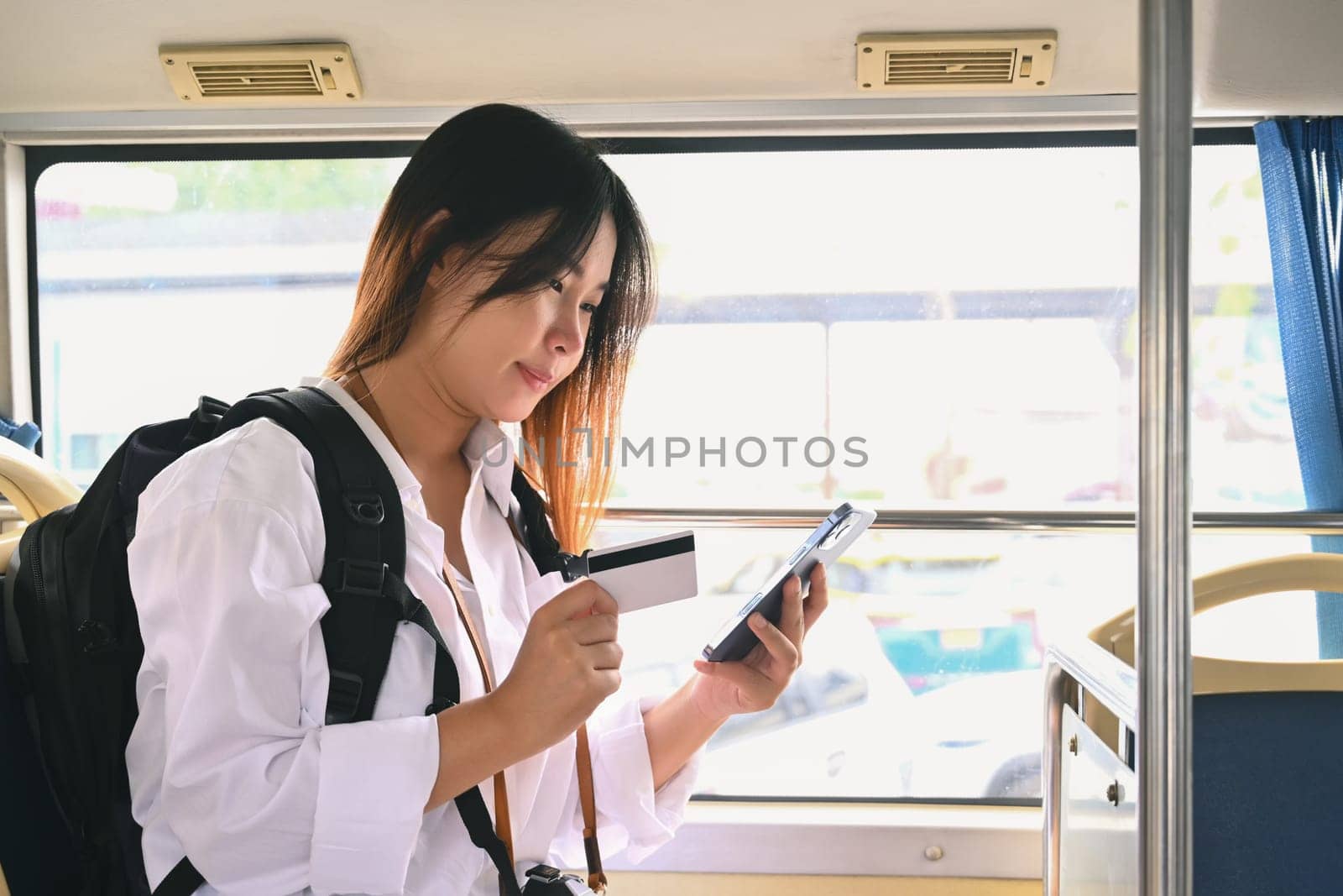 Smiling young woman passenger holding credit card and using mobile phone while traveling by bus.