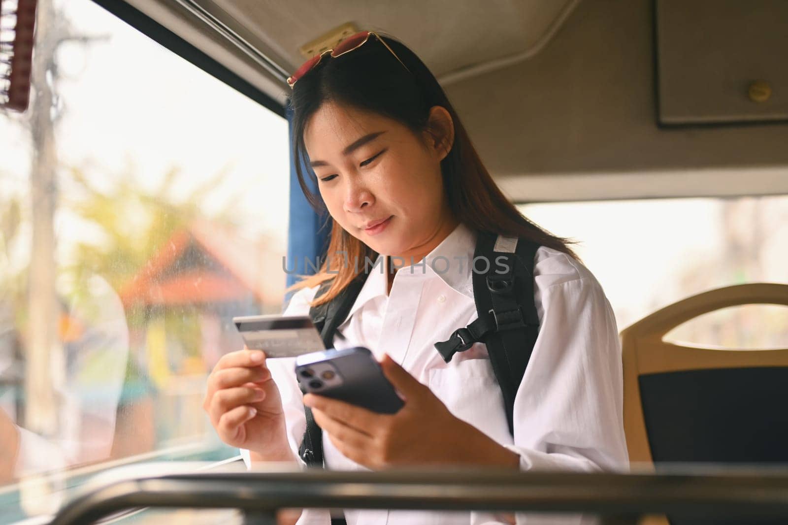 Happy young holding credit card and using mobile phone while sitting inside public bus transport by prathanchorruangsak