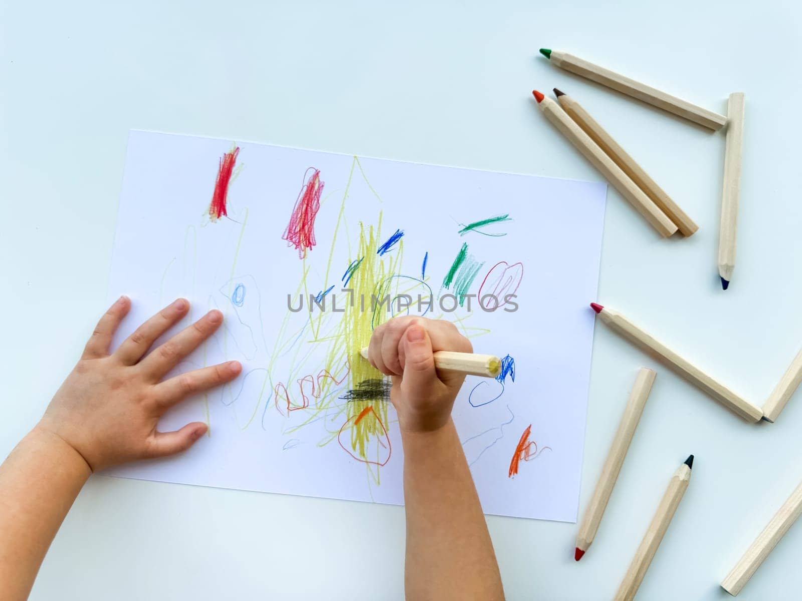 small child draws with colored pencils on paper on white table. High quality photo