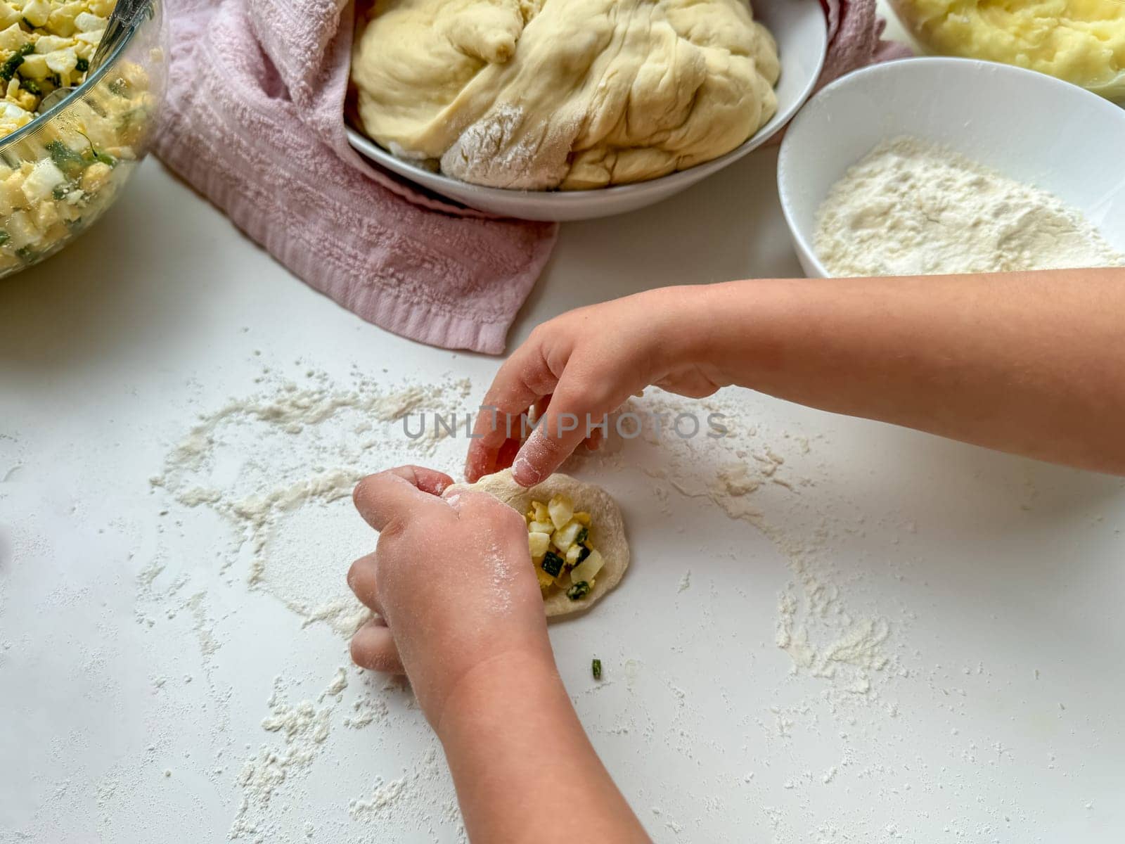 The hands of child knead the dough for making pies on white table, top view. by Lunnica