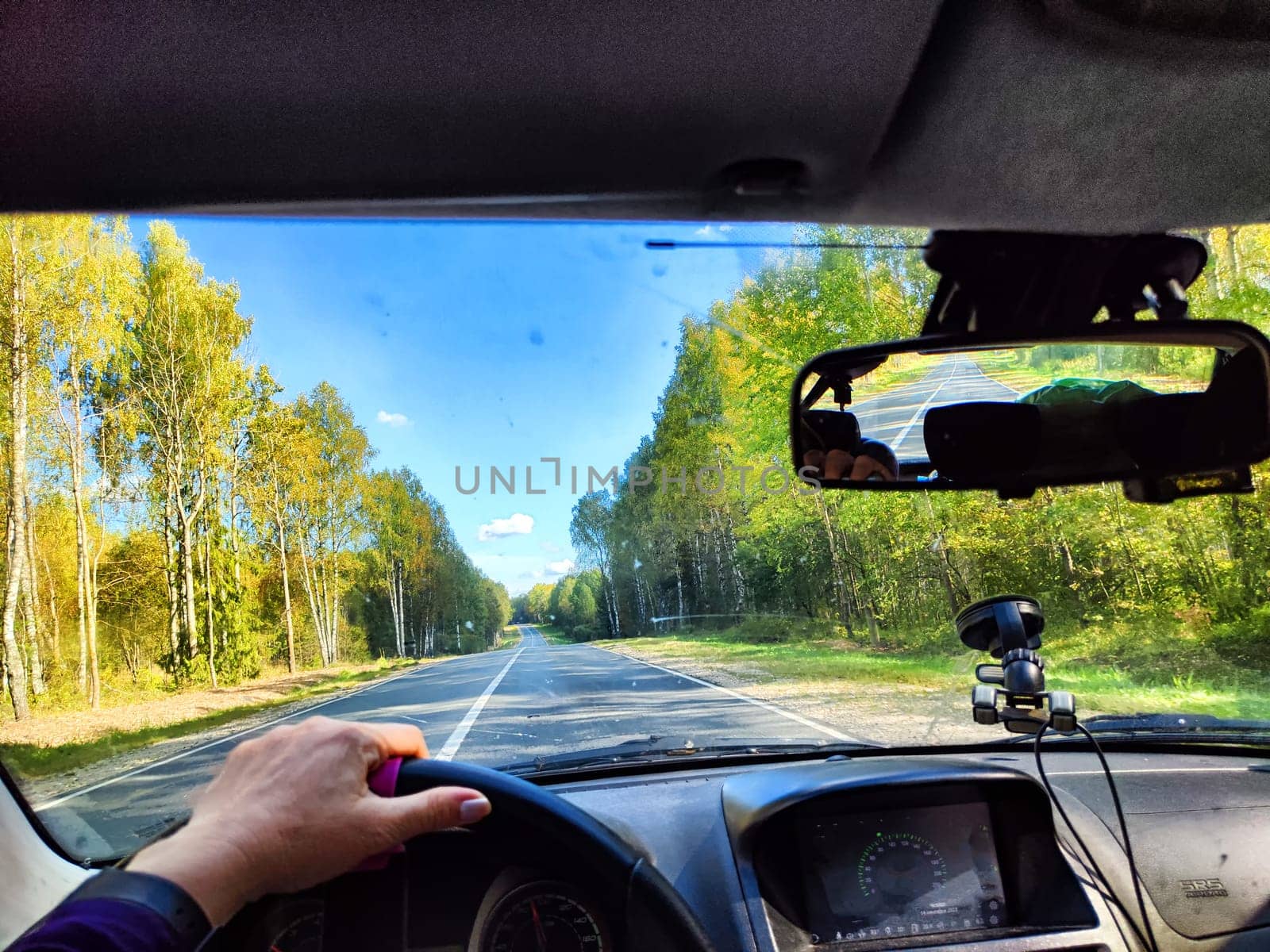 view from a car windshield of natural landscape with road, green trees and blue sky in summer or spring time. Hand of woman on steering wheel. Female traveler driving alone on trip or journey