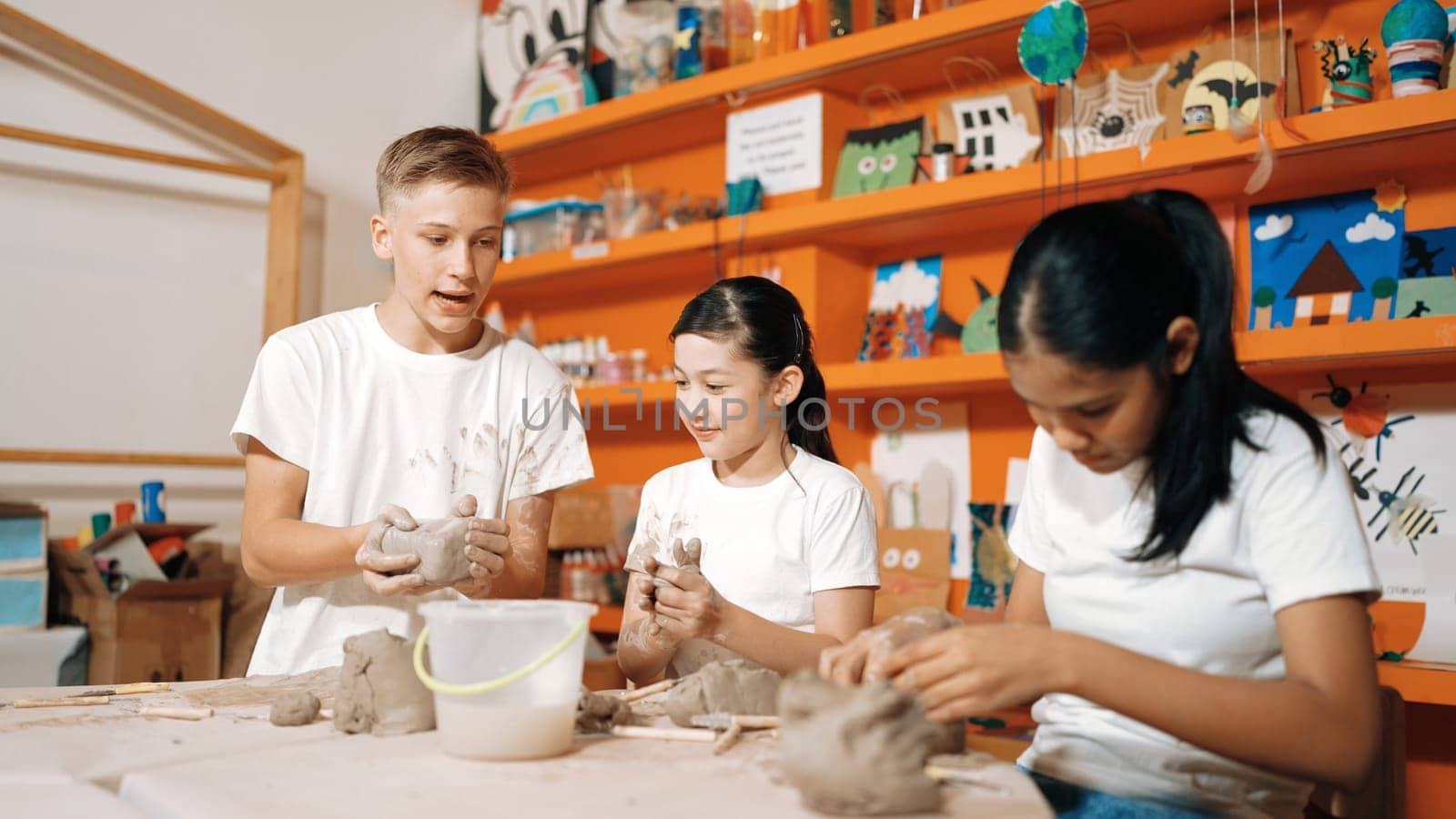 Happy diverse highschool student modeling clay at pottery workshop. Cute children with mixed raced working and making cup of clay by using carving equipment at art lesson. Creative class. Edification.