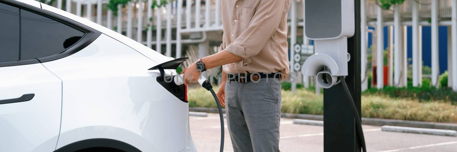 Young man recharge electric car's battery from charging station in city commercial parking lot. Rechargeable EV car for sustainable environmental friendly urban travel. Panorama Expedient