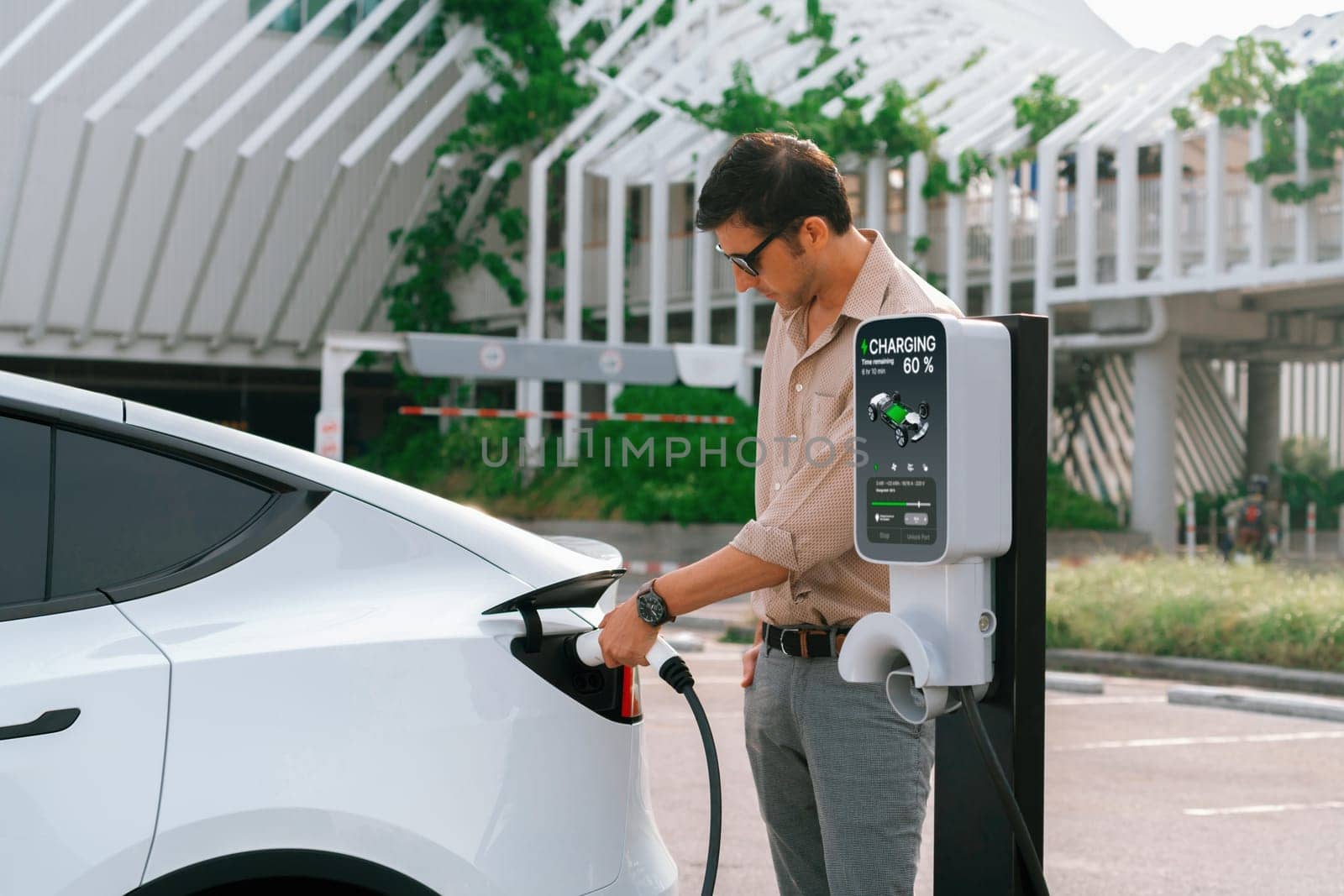 Young man put EV charger to recharge electric car's battery from charging station in city commercial parking lot. Rechargeable EV car for sustainable environmental friendly urban travel. Expedient