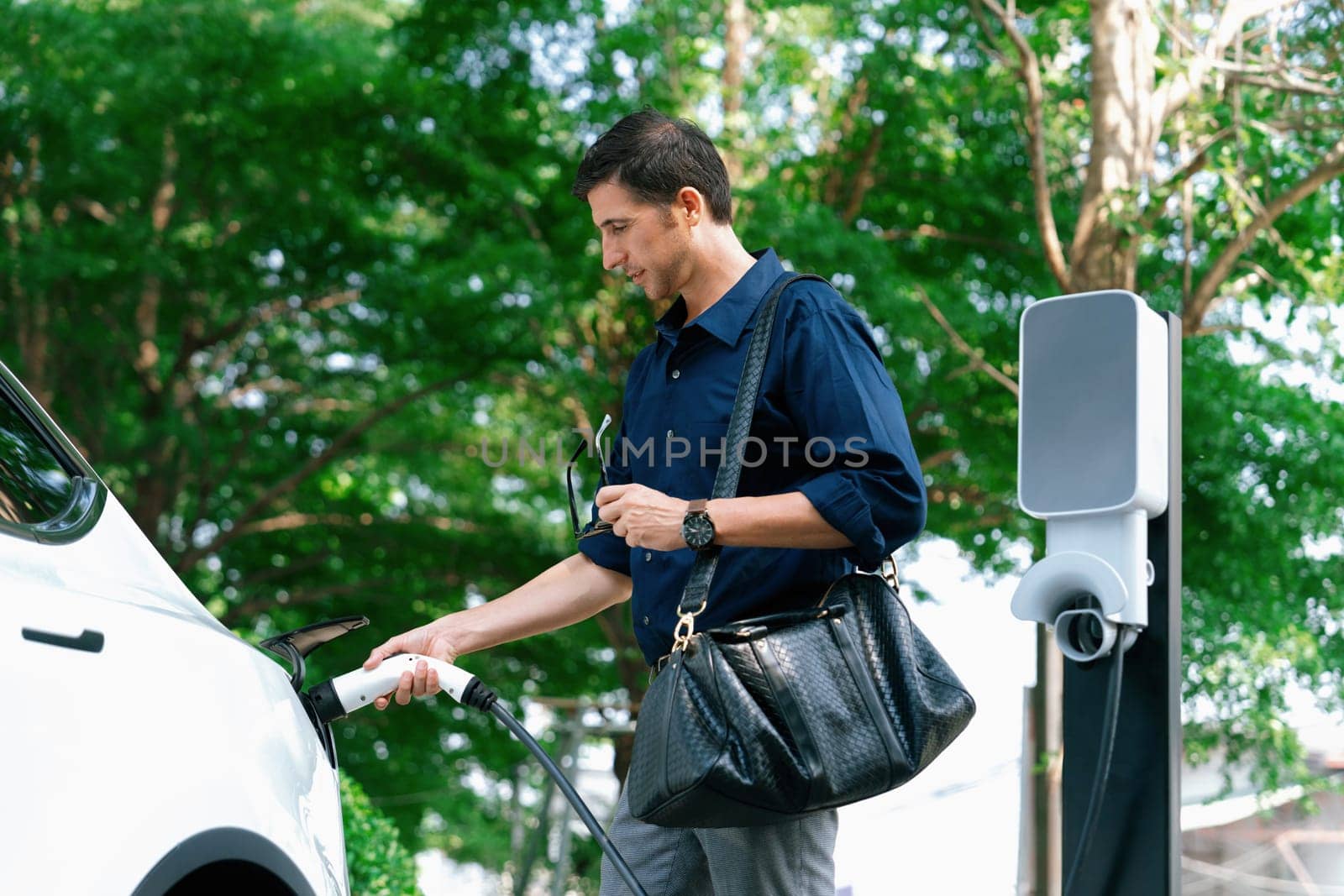 Young man recharge electric car's battery from charging station in outdoor green city park in springtime. Rechargeable EV car for sustainable environmental friendly urban travel lifestyle. Expedient