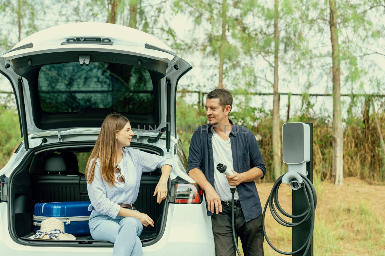 Lovely young couple recharging battery for electric car while relaxing and sitting on the trunk during road trip travel EV car in autumnal forest. Eco friendly travel on vacation during autumn. Exalt