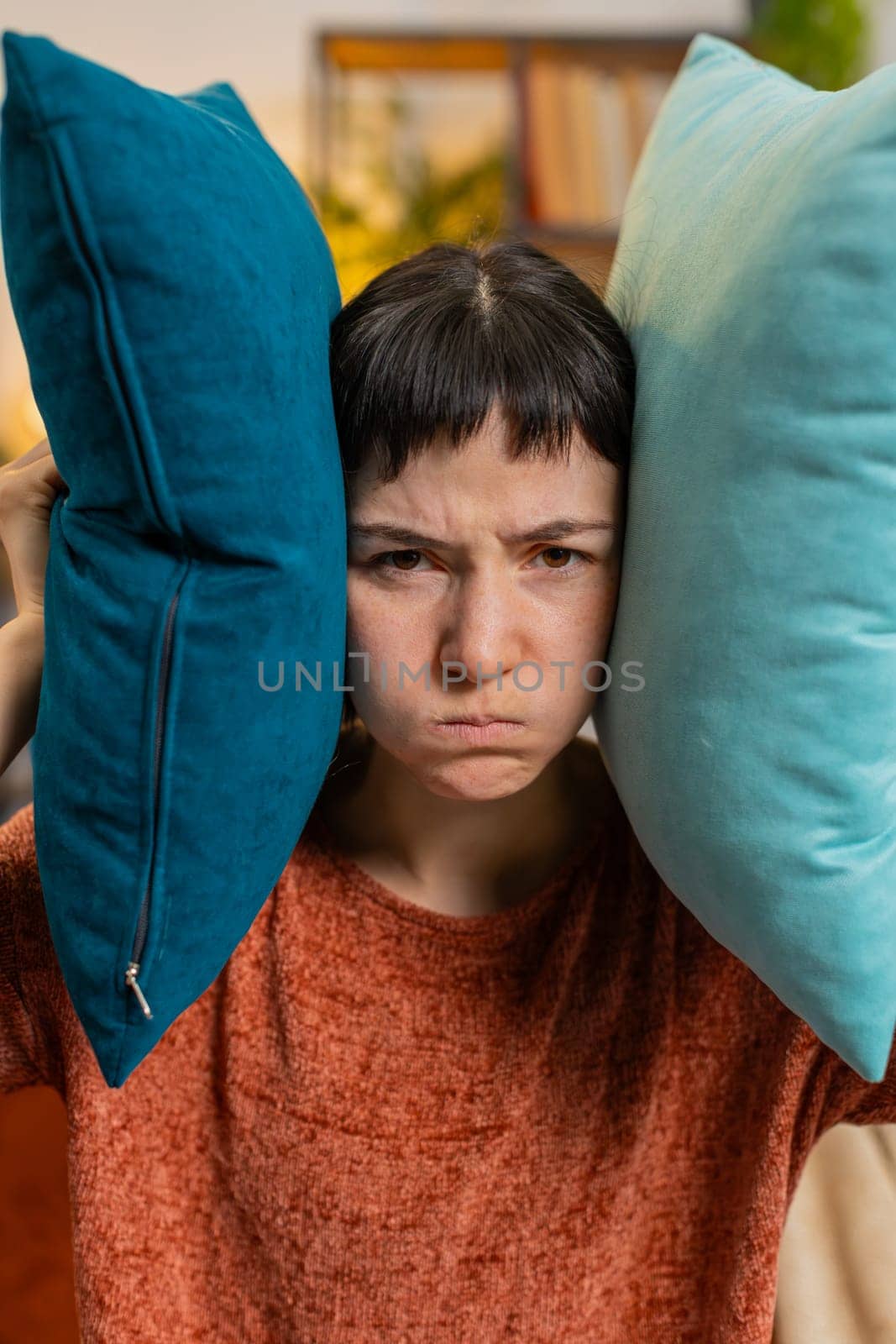 Irritated Caucasian woman girl covers ears with cushions, annoyed by noisy neighbors suffers from headache sitting on sofa. Thin walls at home, flat without sound insulation, repair work concept