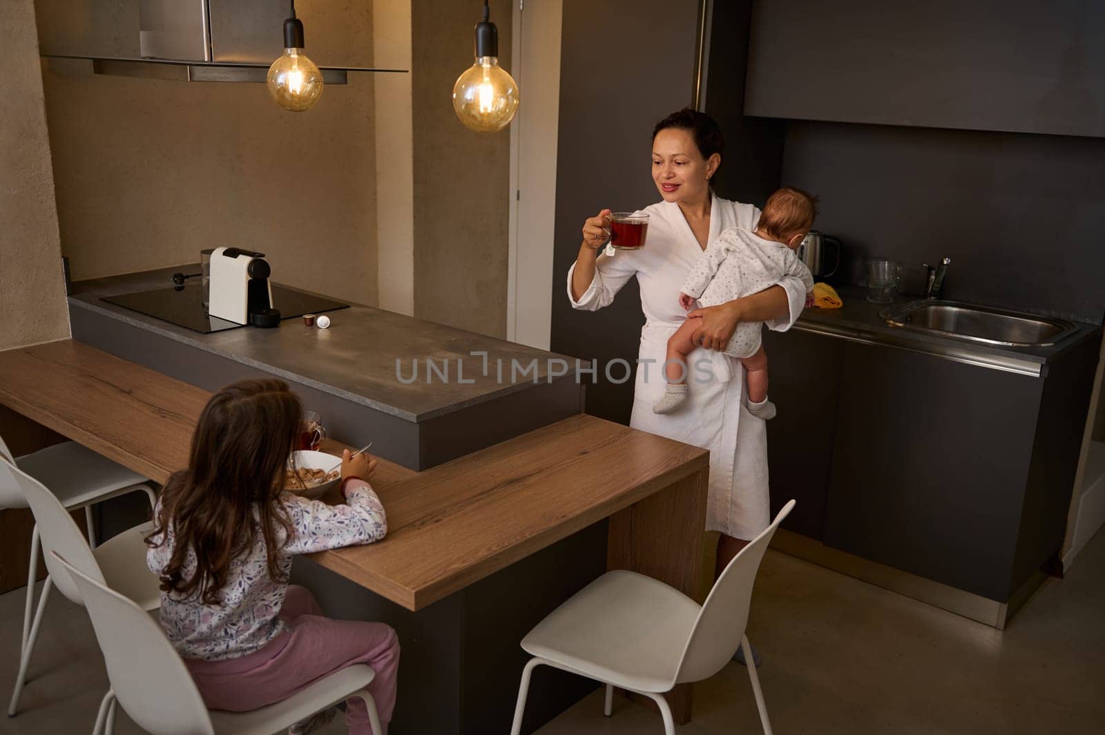 Portrait of young happy family of a loving mother carrying her newborn baby, drinking hot tea and enjoying a nice conversation with her daughter who is taking breakfast in the morning while waking up