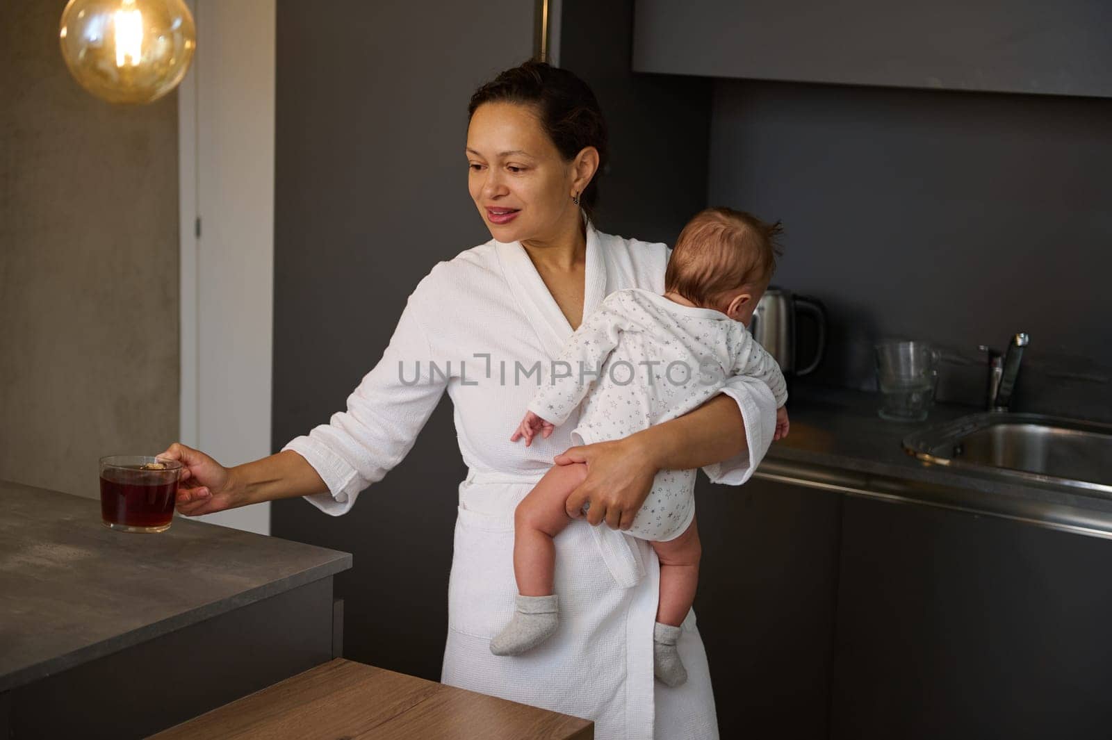 Beautiful multi ethnic young woman in white bathrobe, a loving happy carrying her baby and drinking tea in the morning for while breakfast time. People. Food and drink. Maternity lifestyle