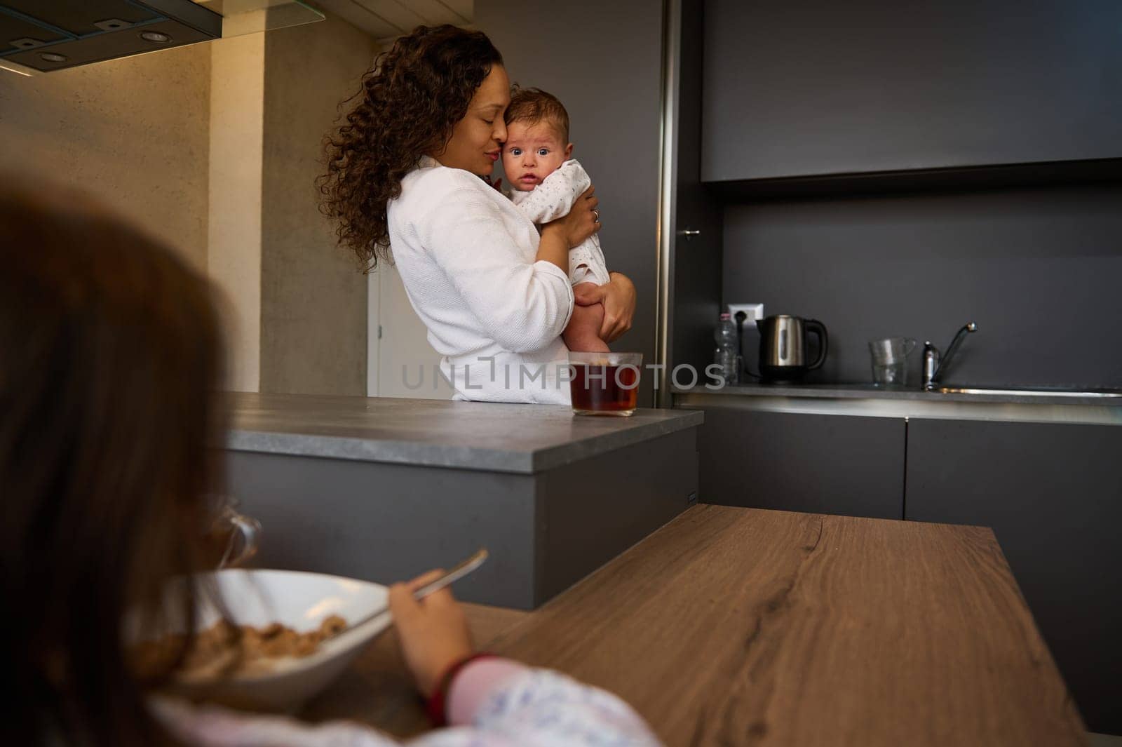 Delightful mother gently hugging her baby boy, standing together in the kitchen while older child daughter taking her breakfast, experiencing jealousy looking at her mom carrying her little brother