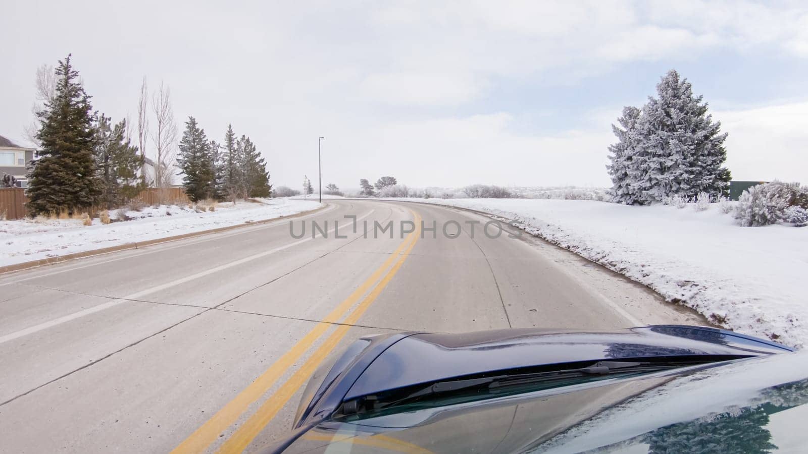 Traversing a freshly cleared, suburban road after a winter storm, one experiences a serene drive through an upscale residential neighborhood. Snow-covered houses and trees contribute to a picturesque winter scene, enhancing the tranquility of the journey.
